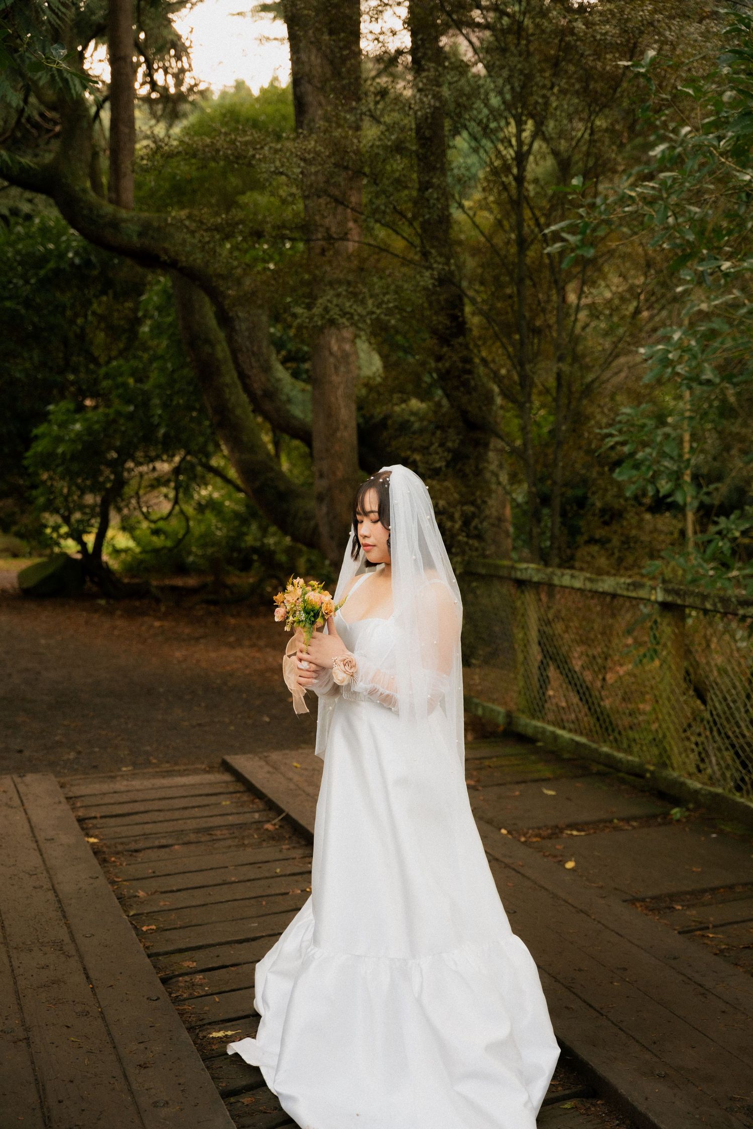Bride in white gown and veil holds bouquet on wooden path surrounded by lush green trees.