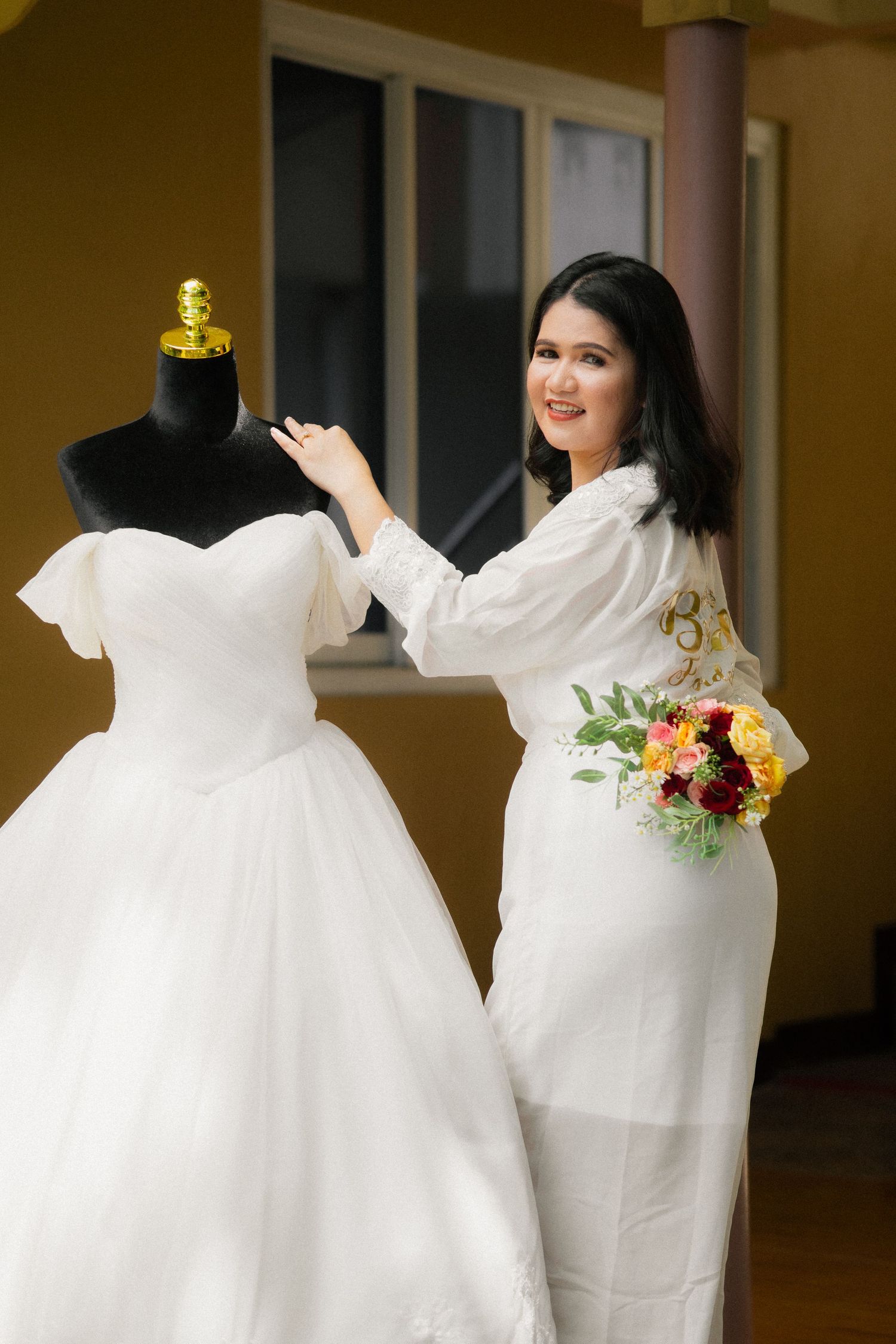 Woman in white dress holding flowers beside a bridal gown on a mannequin, smiling in a bright room.