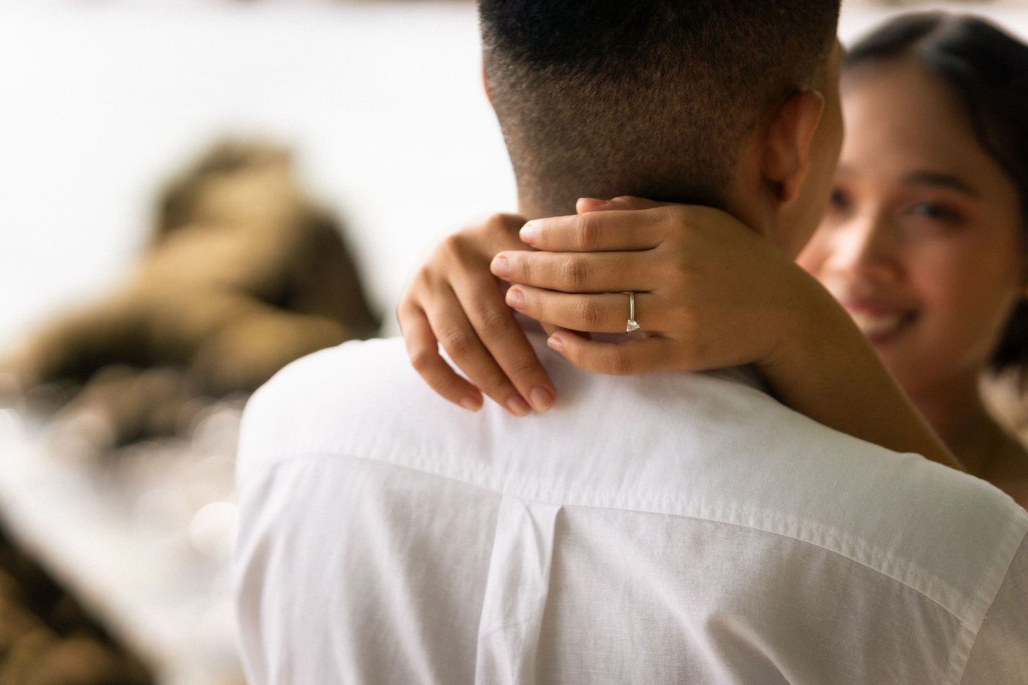 A woman with an engagement ring embraces a man in a white shirt in a close romantic moment.