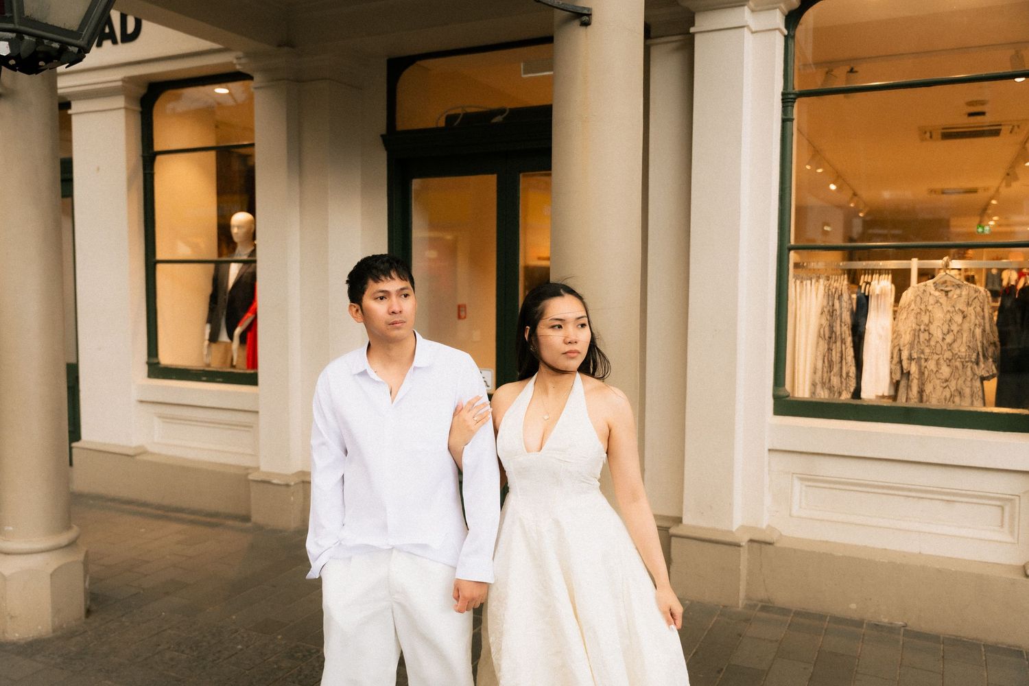 A couple in white outfits poses in front of an upscale boutique store, woman in halter dress, man in casual shirt.