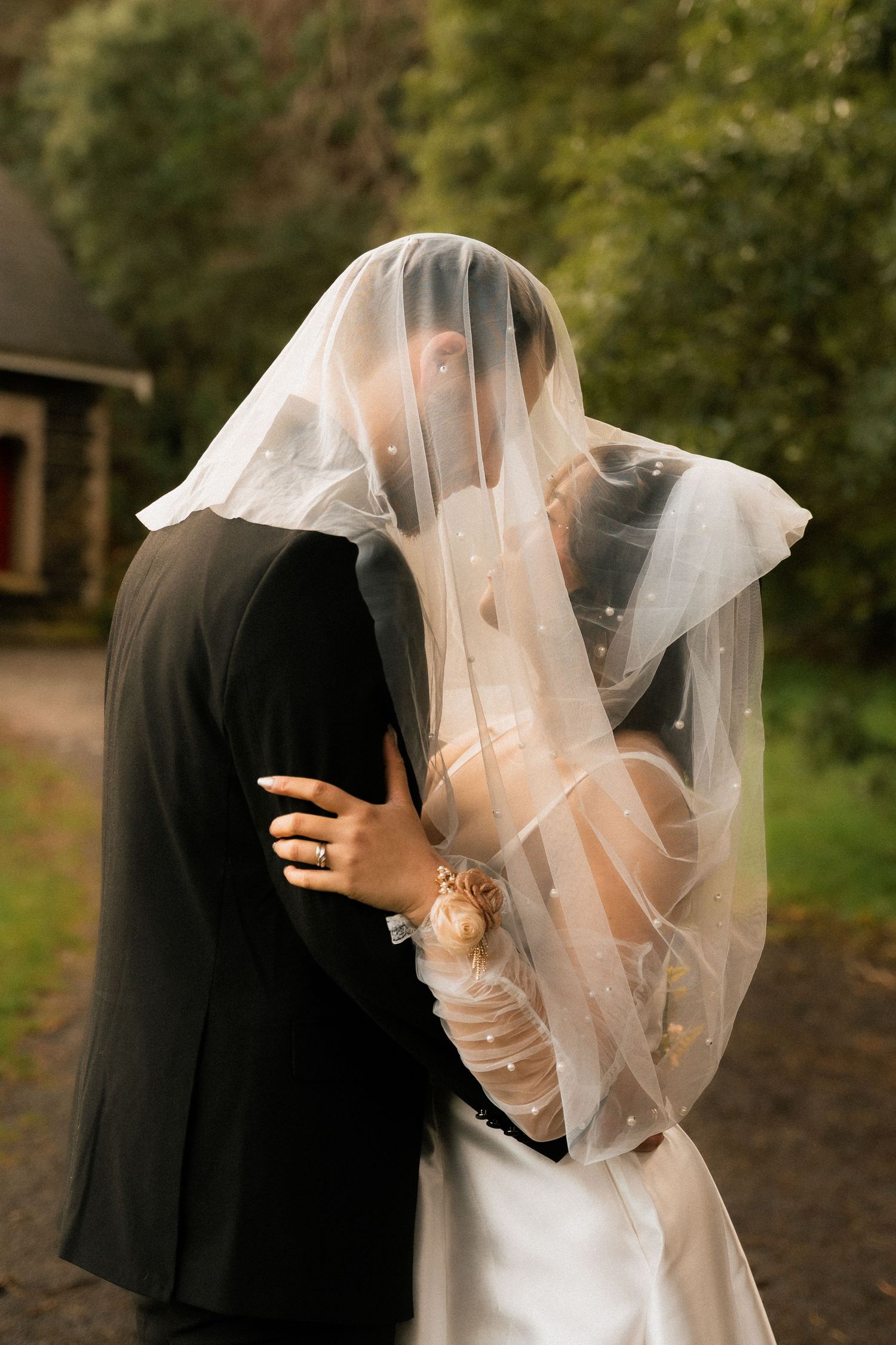 Bride and groom kissing under a sheer wedding veil outdoors, surrounded by lush green trees.
