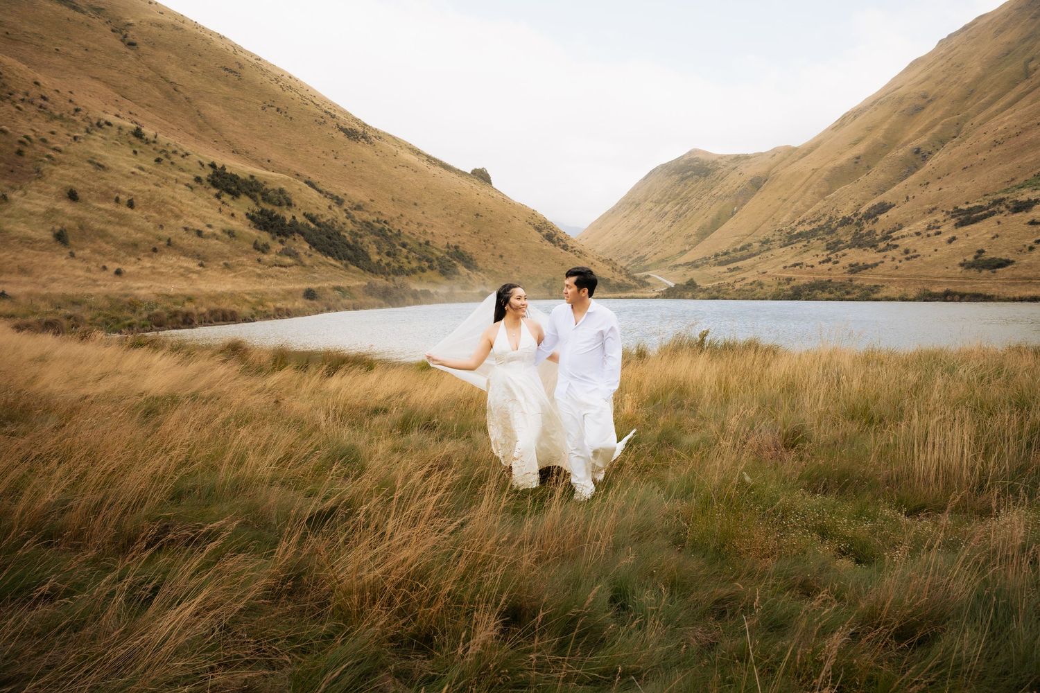 Couple in white wedding attire walking through golden grass fields with a mountain lake backdrop in New Zealand.