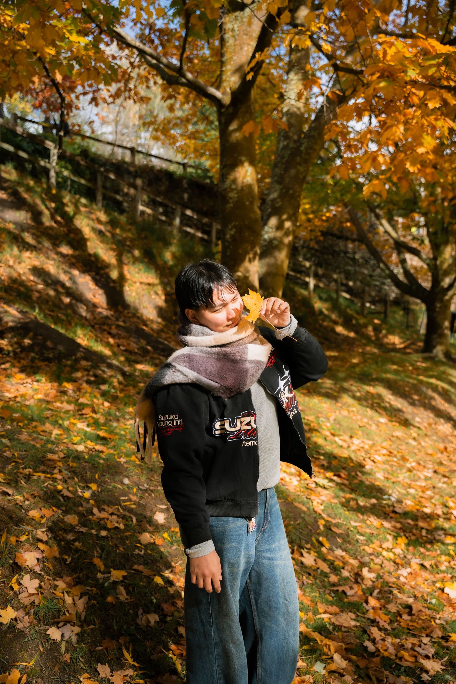 Person holding autumn leaf in fall park with golden maple trees and scattered leaves on ground.
