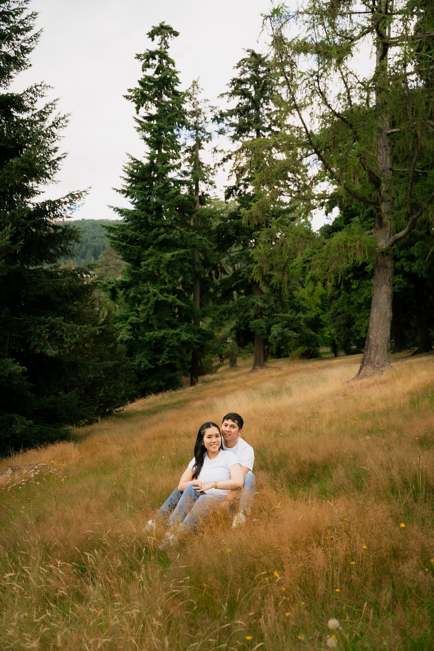 Couple sitting together in golden meadow grass surrounded by tall evergreen trees on a forested hillside.