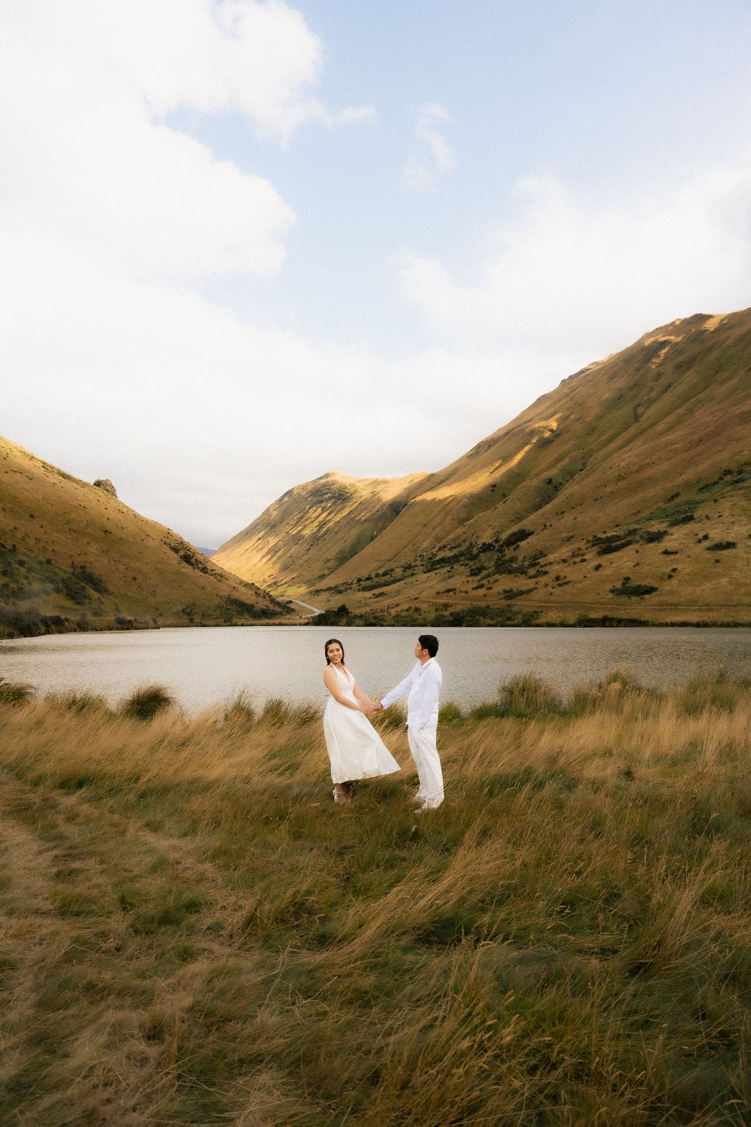 Couple in white holding hands in golden grass field with mountain lake backdrop during outdoor engagement shoot.