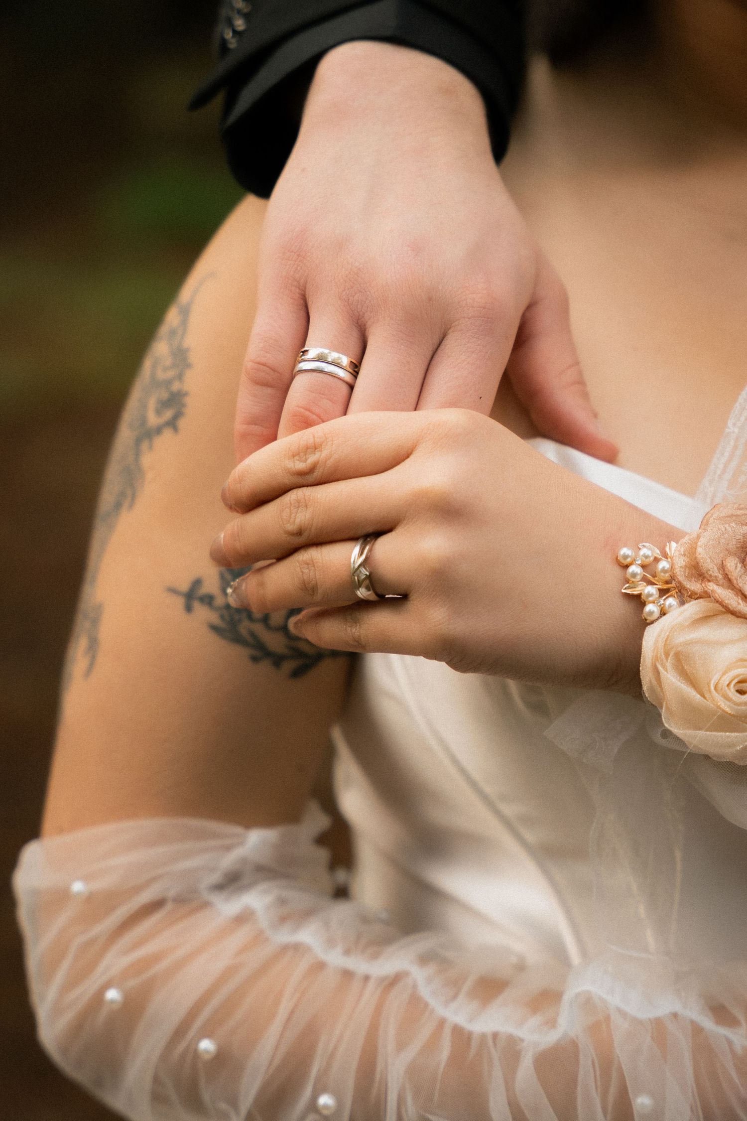 Close-up of bride and groom hands with wedding rings, tattoos, pearl bracelet, and sheer white dress sleeve.