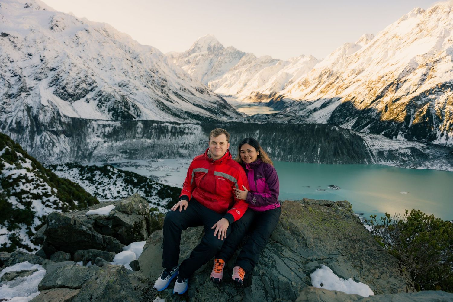 Smiling couple posed on rocky outcrop with panoramic view of snowy mountains and glacial lake below.