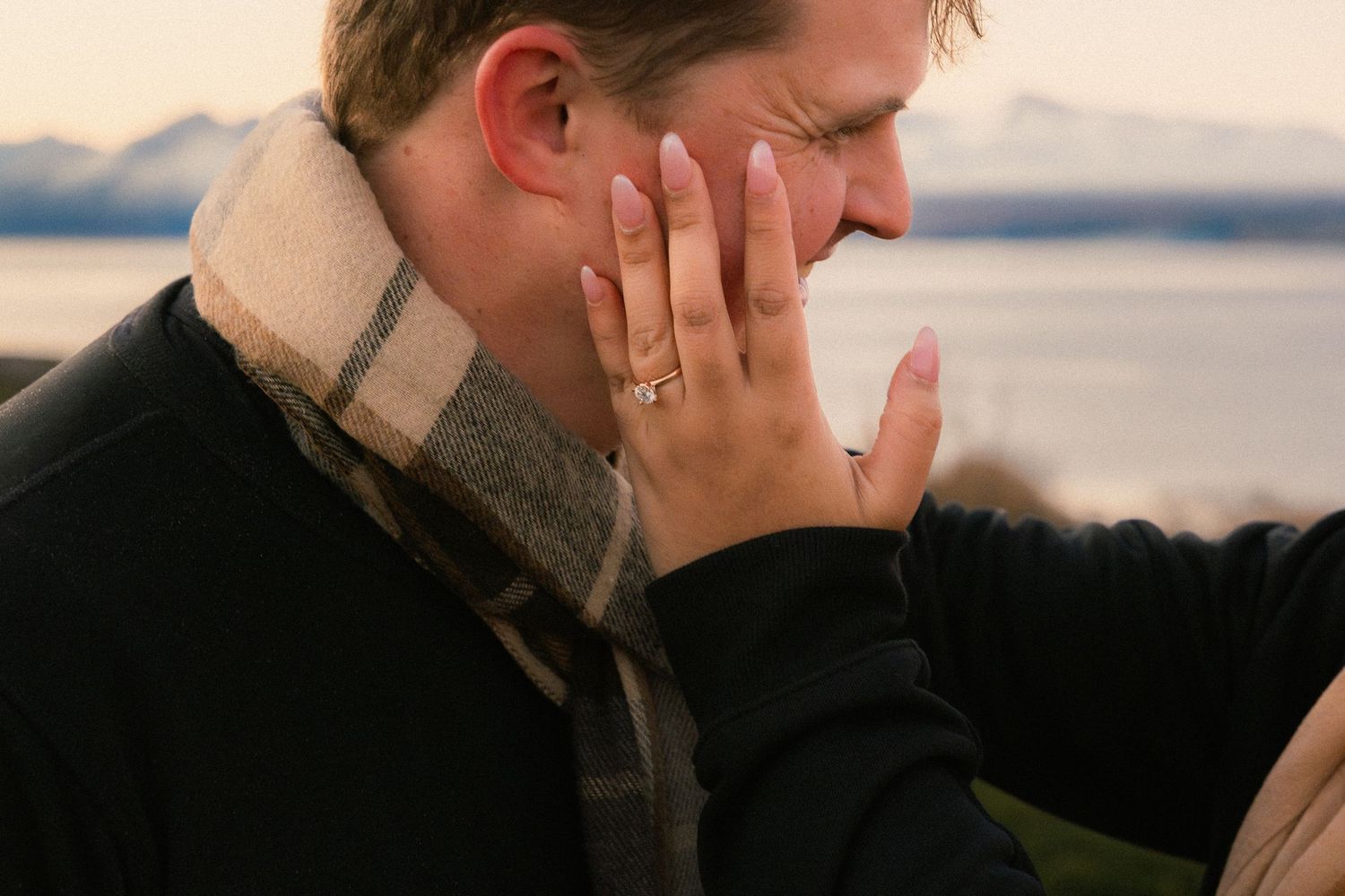 Couple sharing a tender moment outdoors, woman wearing engagement ring touching man's face near waterfront.