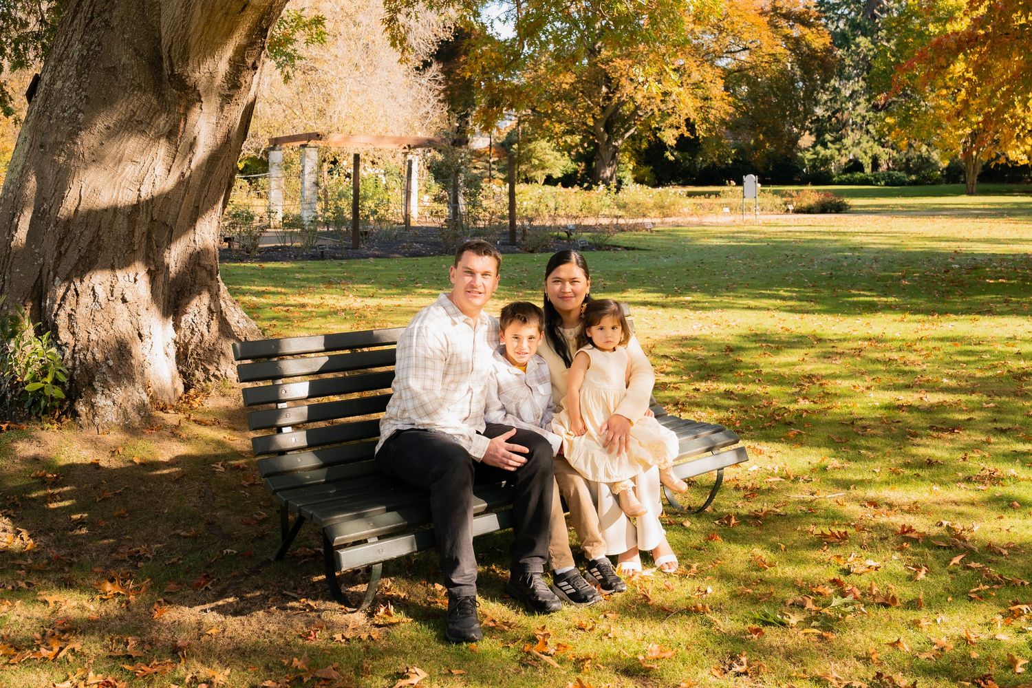 Family of four sitting on a park bench surrounded by autumn foliage and golden trees on a sunny day.