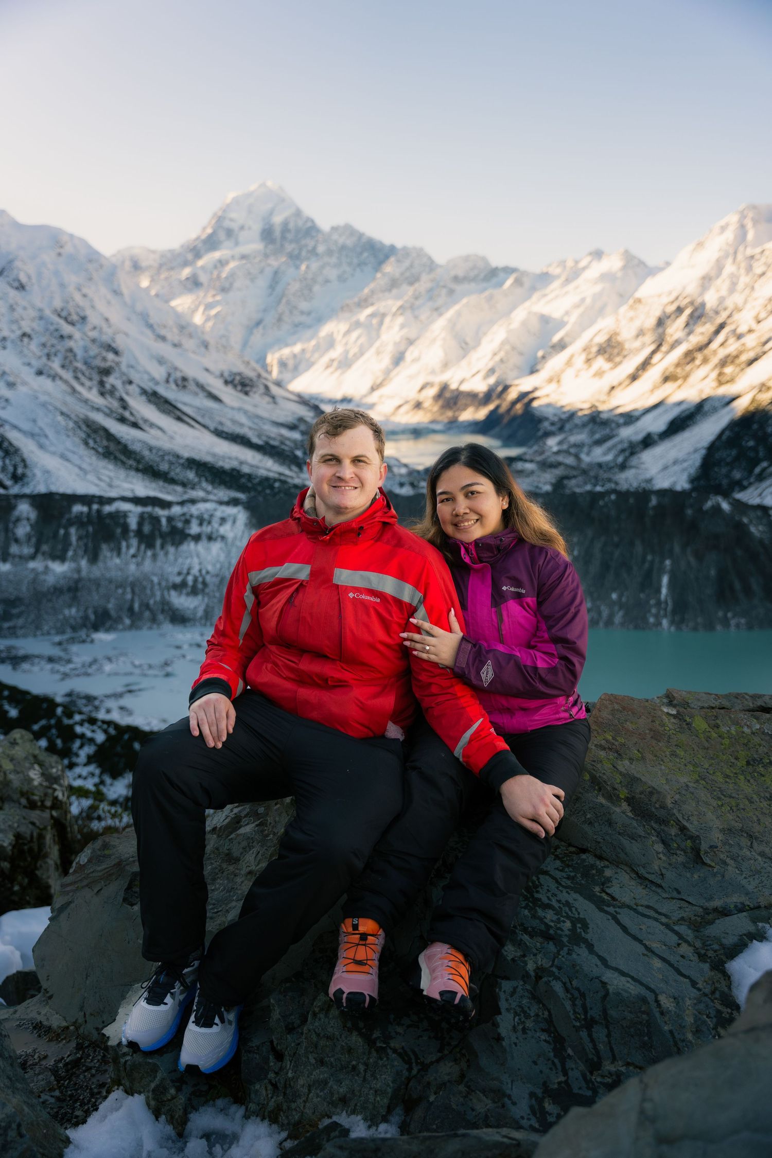 Happy couple sitting on rocks with stunning snow-capped mountains and turquoise glacial lake backdrop.
