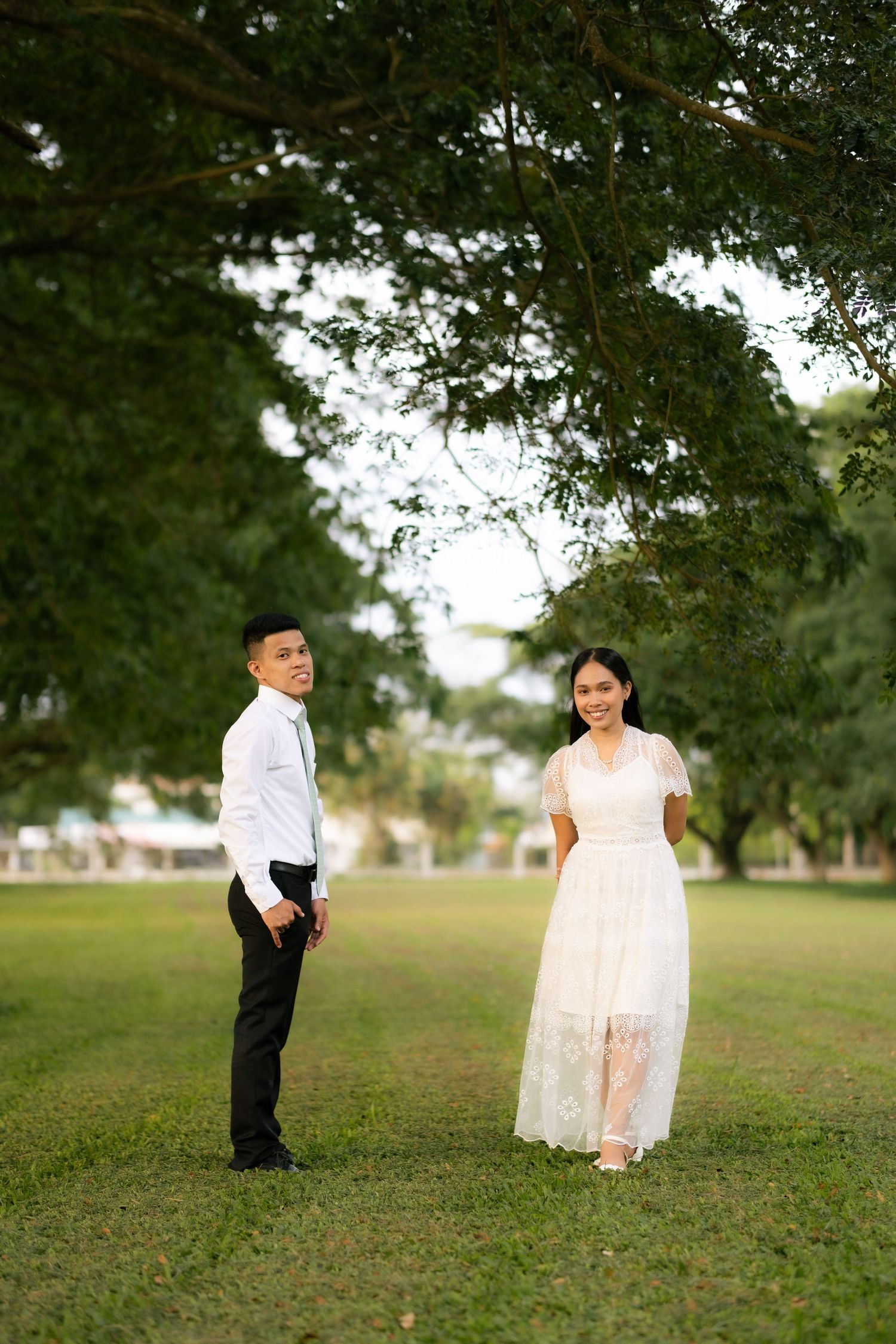 A couple in formal wear stands apart on a green lawn beneath large shady trees smiling at the camera.