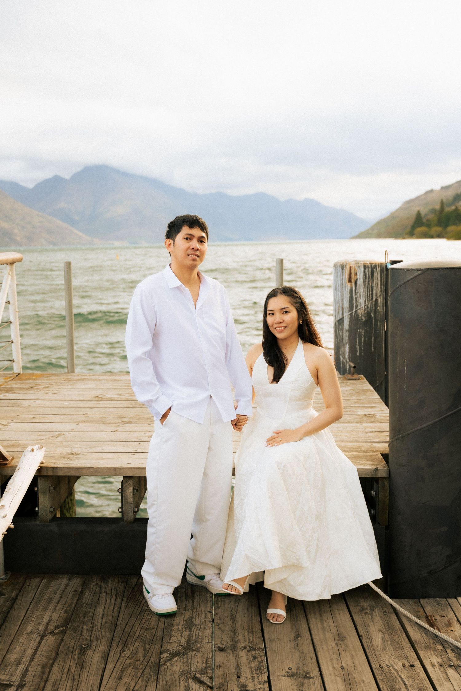 Couple in white outfits posing on a wooden dock with mountain lake backdrop.