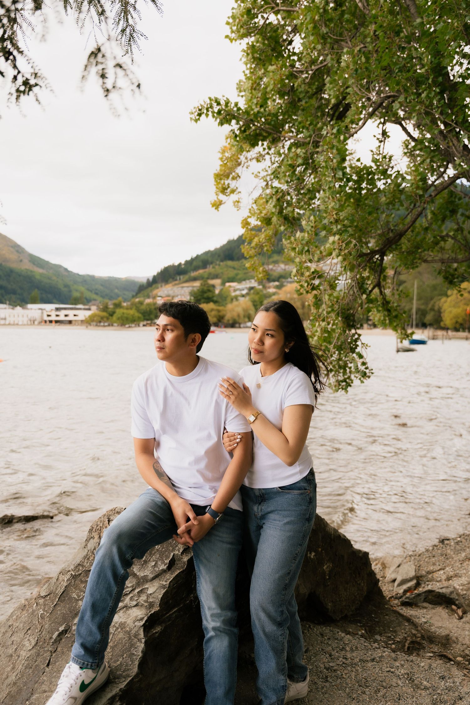 A couple in matching white t-shirts and jeans sit on rocks by a lakeside, with green trees and mountains in the background.