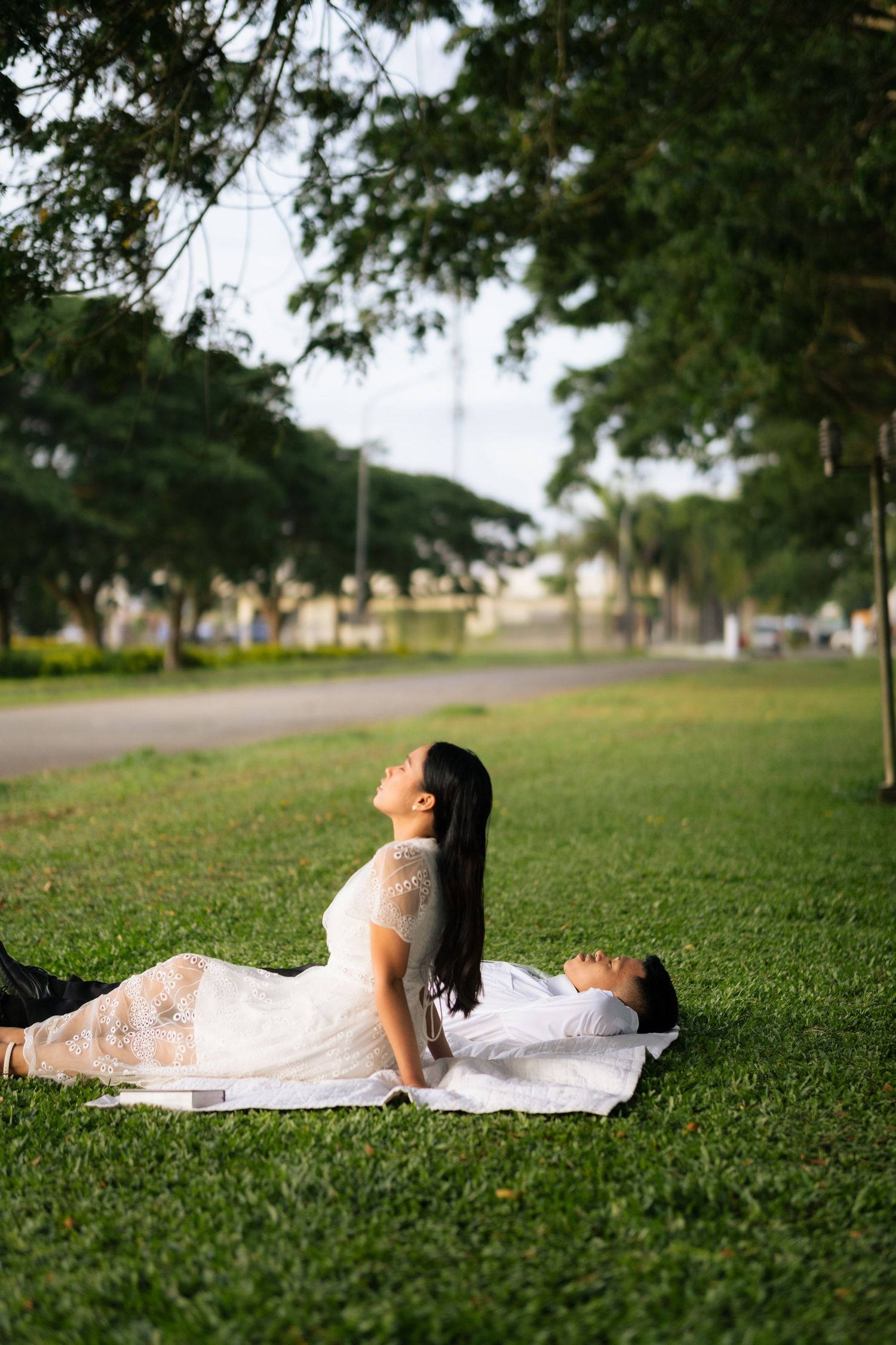 A woman in a white lace dress sits on grass while her partner lies beside her in a lush green park.