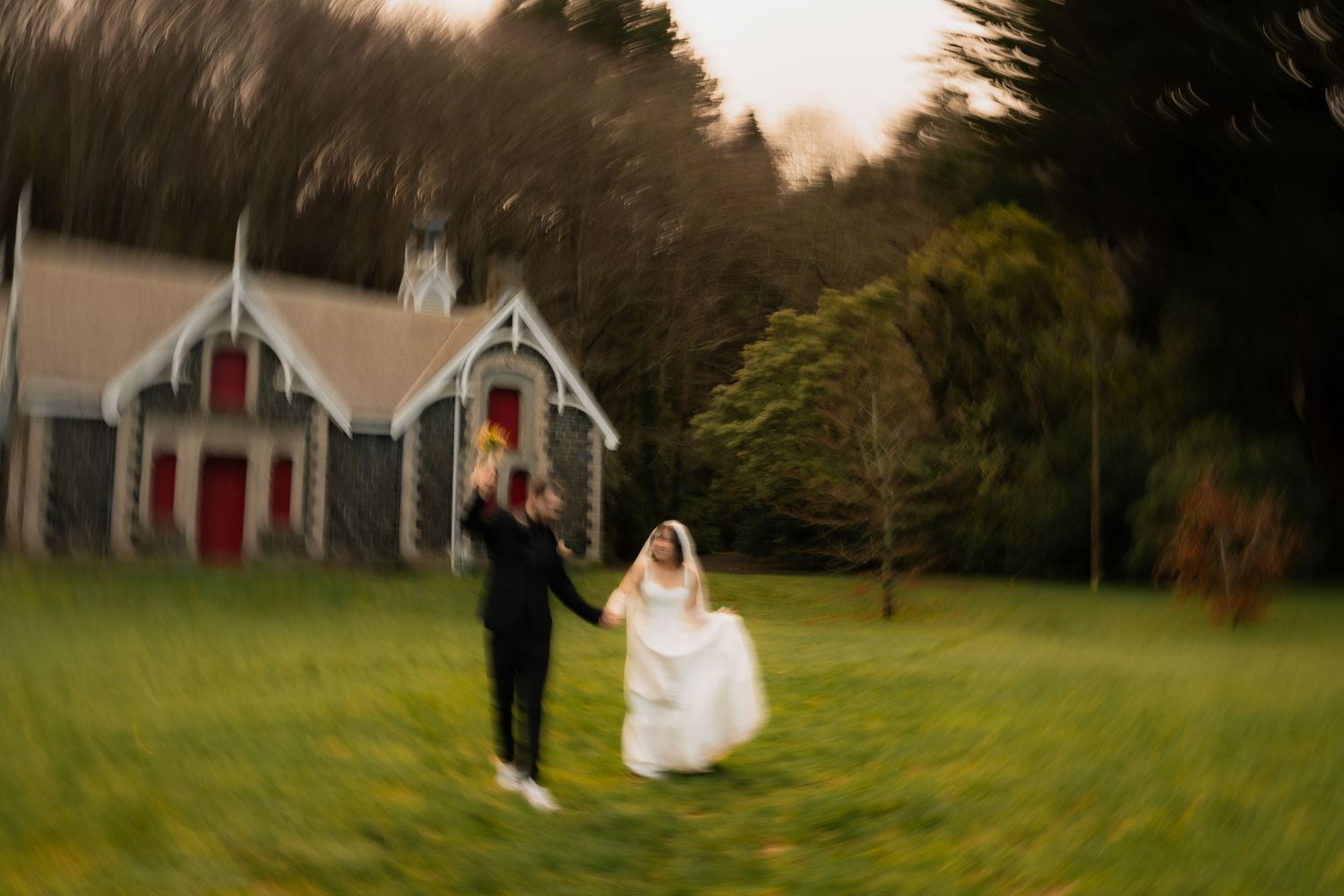 Bride and groom holding hands on green lawn outside rustic stone chapel surrounded by trees.