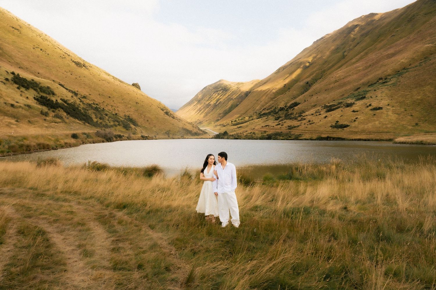 Couple in white outfits embracing by a serene mountain lake with golden grass fields in New Zealand.