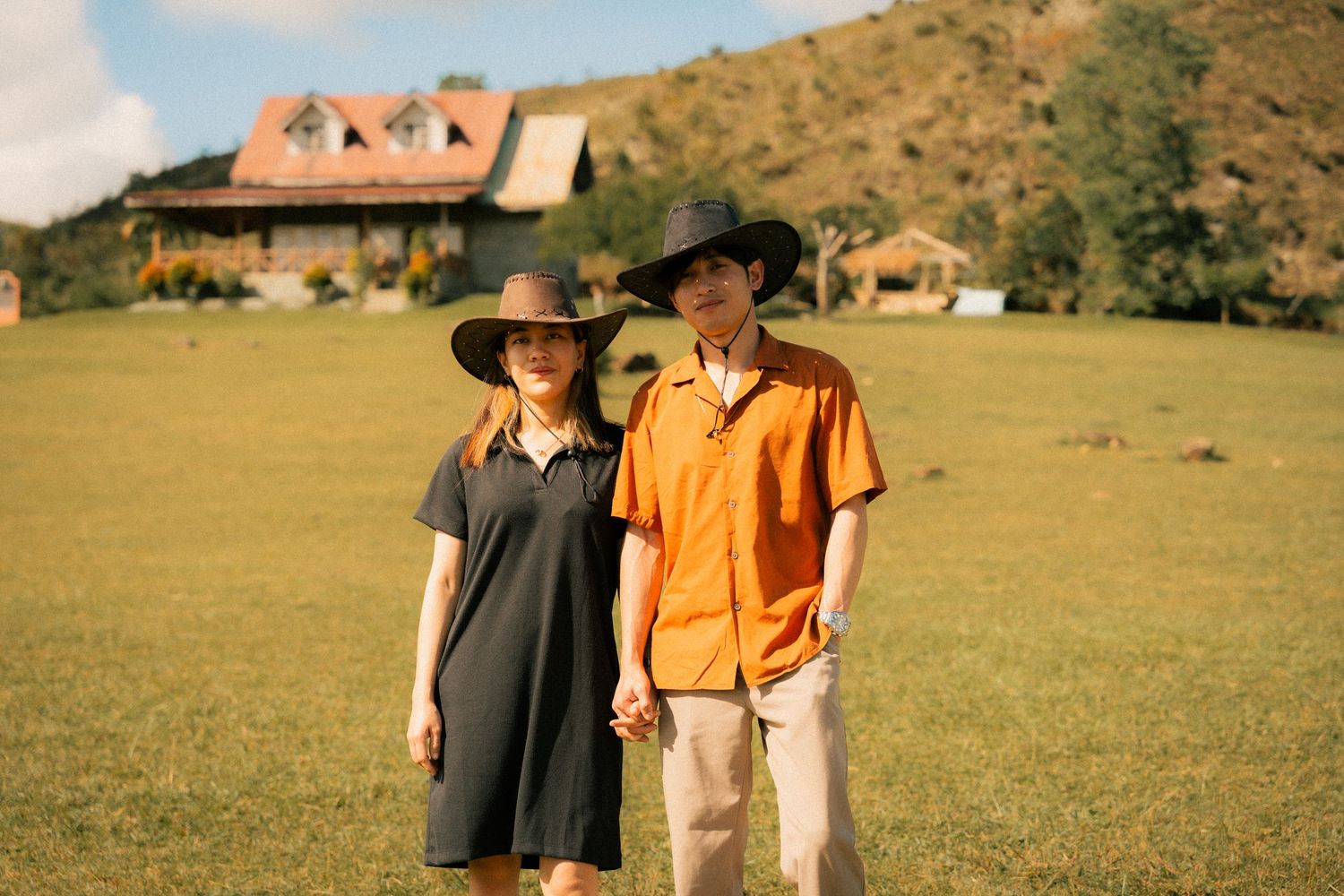 Couple in wide-brim hats holding hands on green field, woman in black dress, man in orange shirt, rustic house behind.