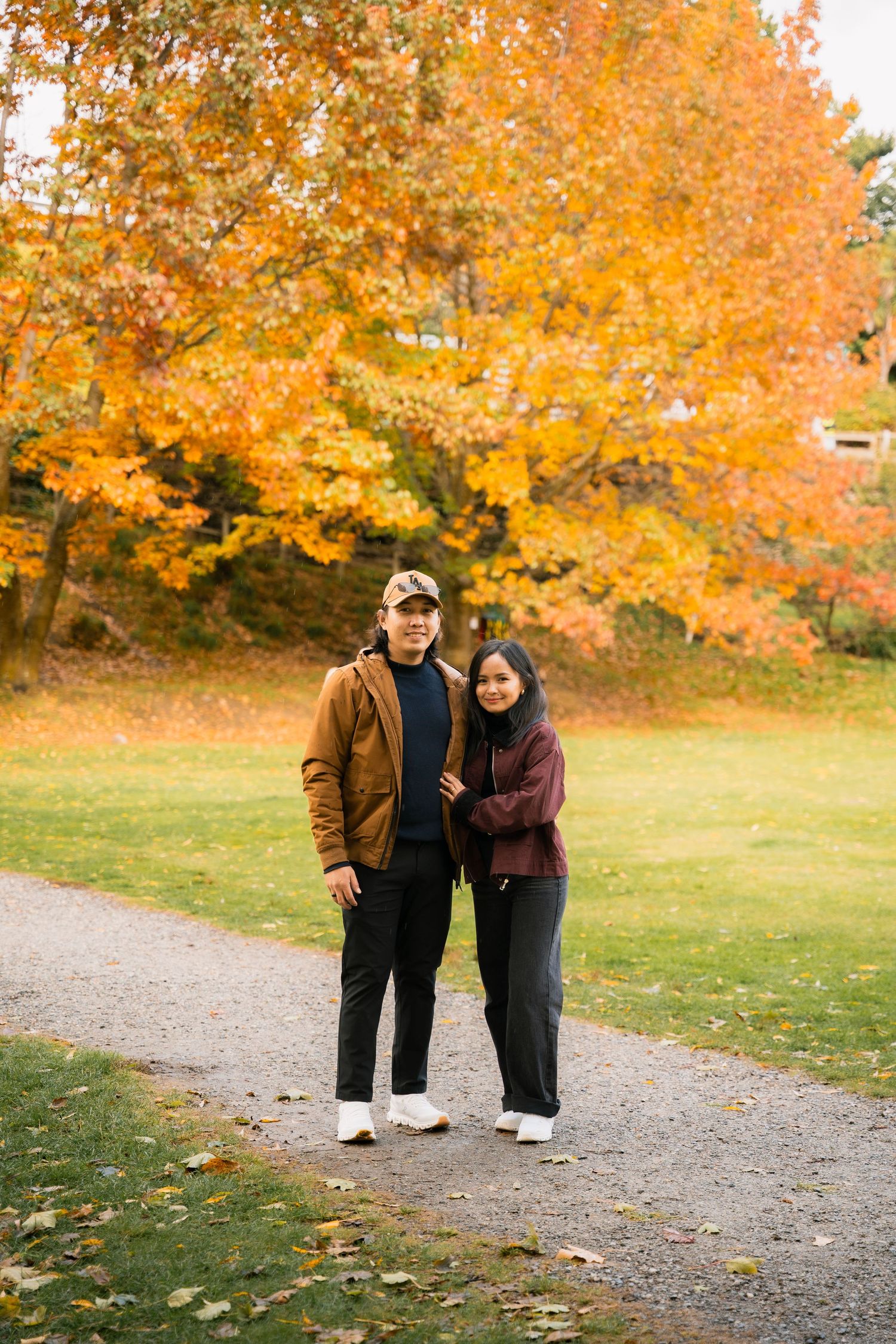 A couple poses on a gravel path surrounded by vibrant orange autumn foliage in a scenic park setting.