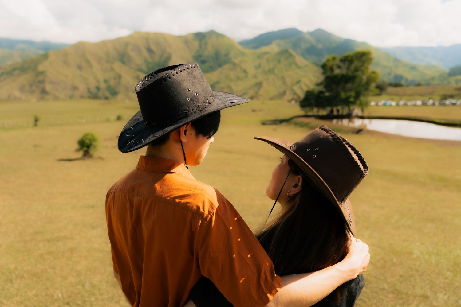 Couple in cowboy hats embracing while admiring scenic mountain landscape on a sunny day.