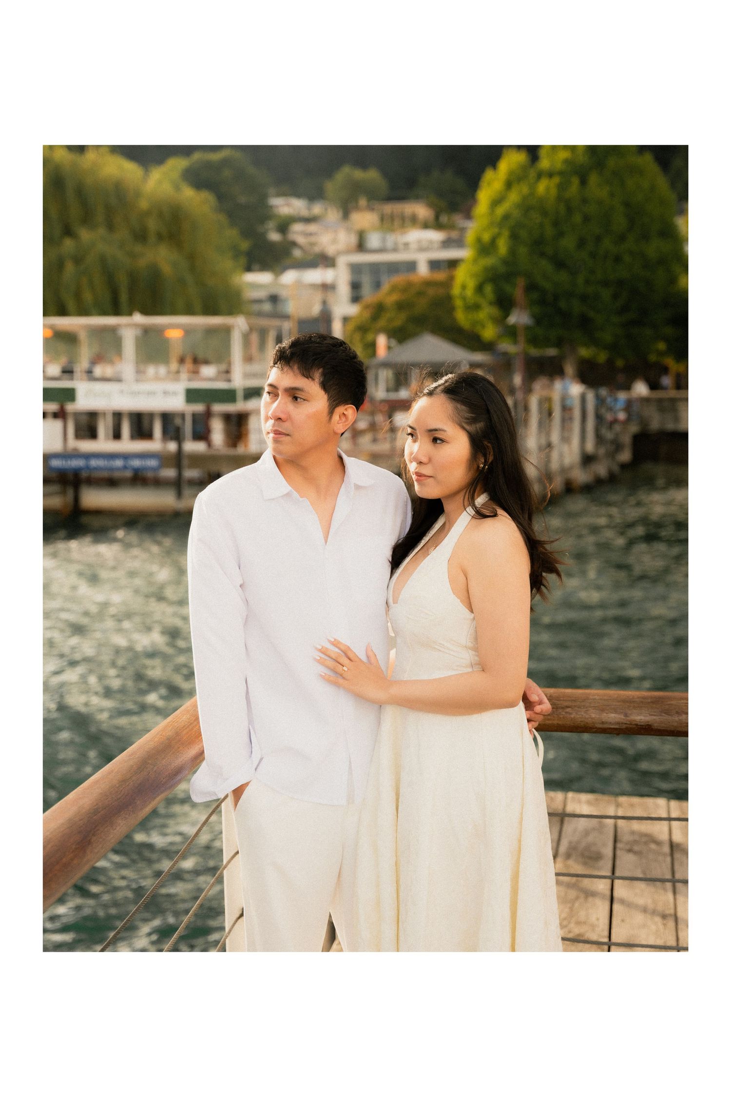 Couple in white outfits posing on a waterfront dock with trees and buildings in the background.