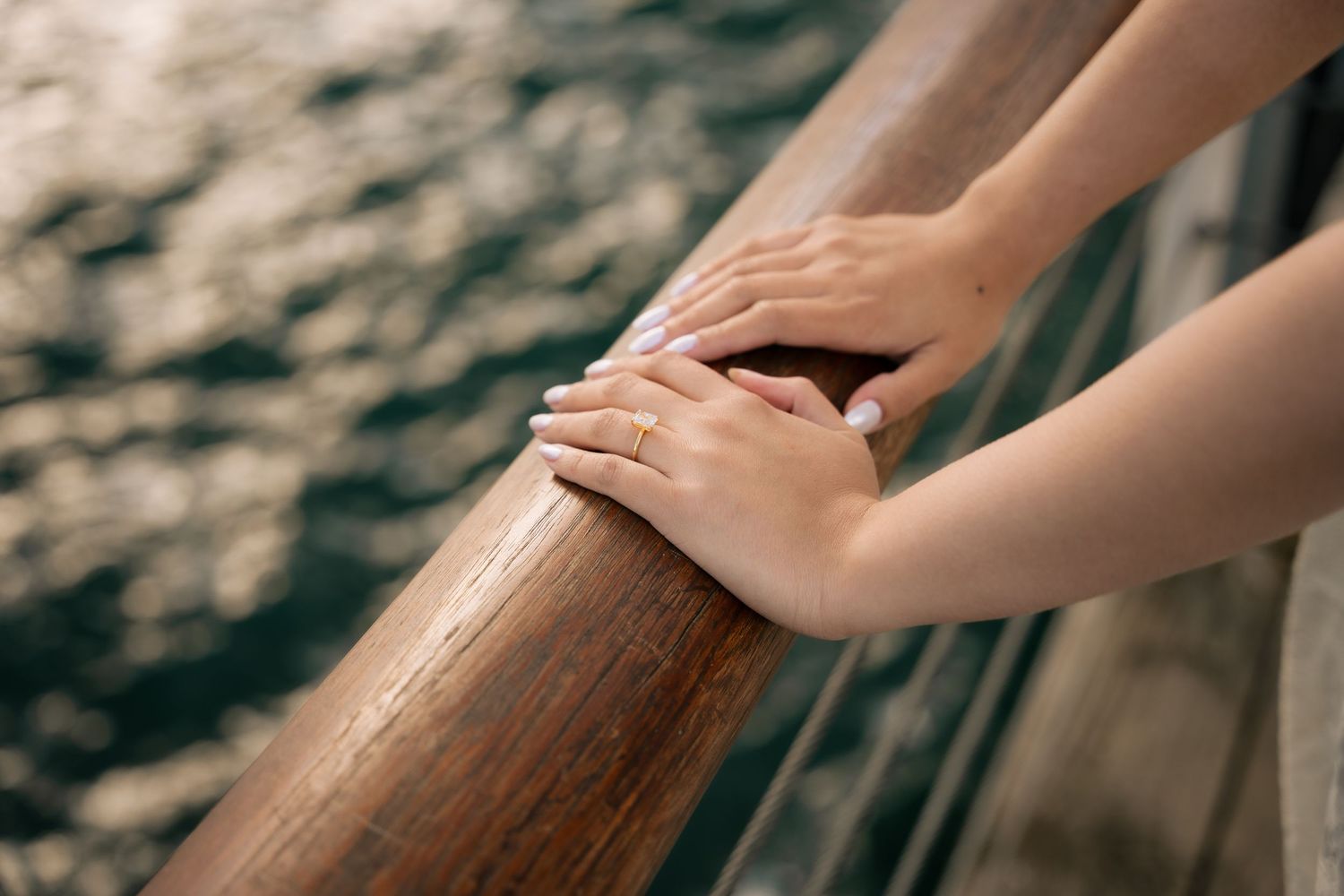 Two hands resting on a wooden railing overlooking shimmering water on a cruise ship deck.