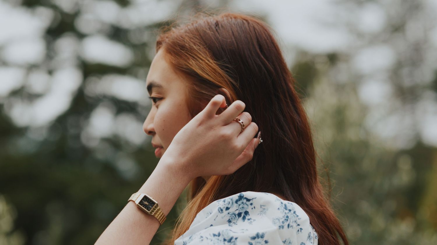 Young woman with auburn hair adjusting wireless earbud, wearing a floral blouse outdoors.