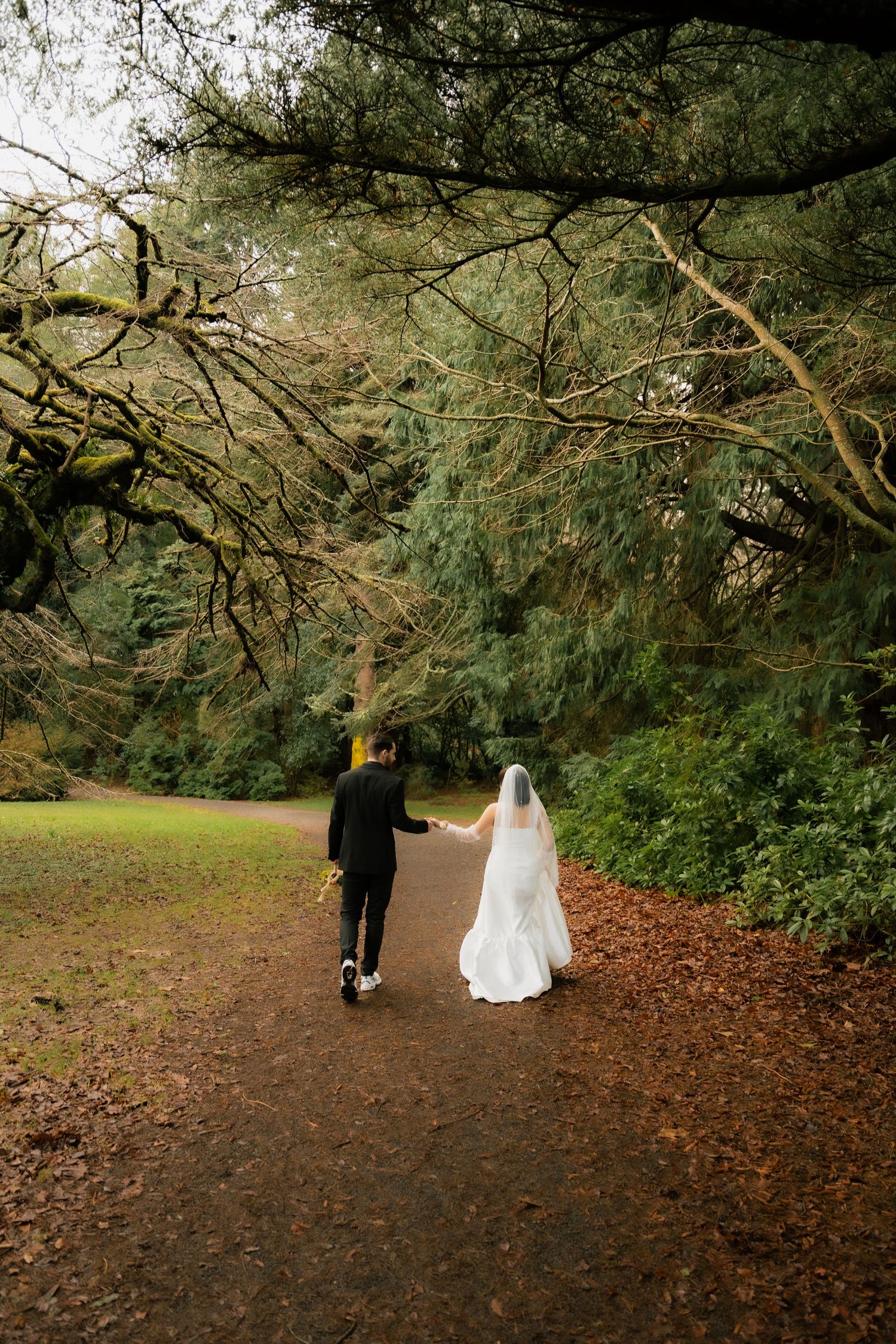 A bride and groom walk hand in hand along a forest path, her white gown and veil trailing behind them.