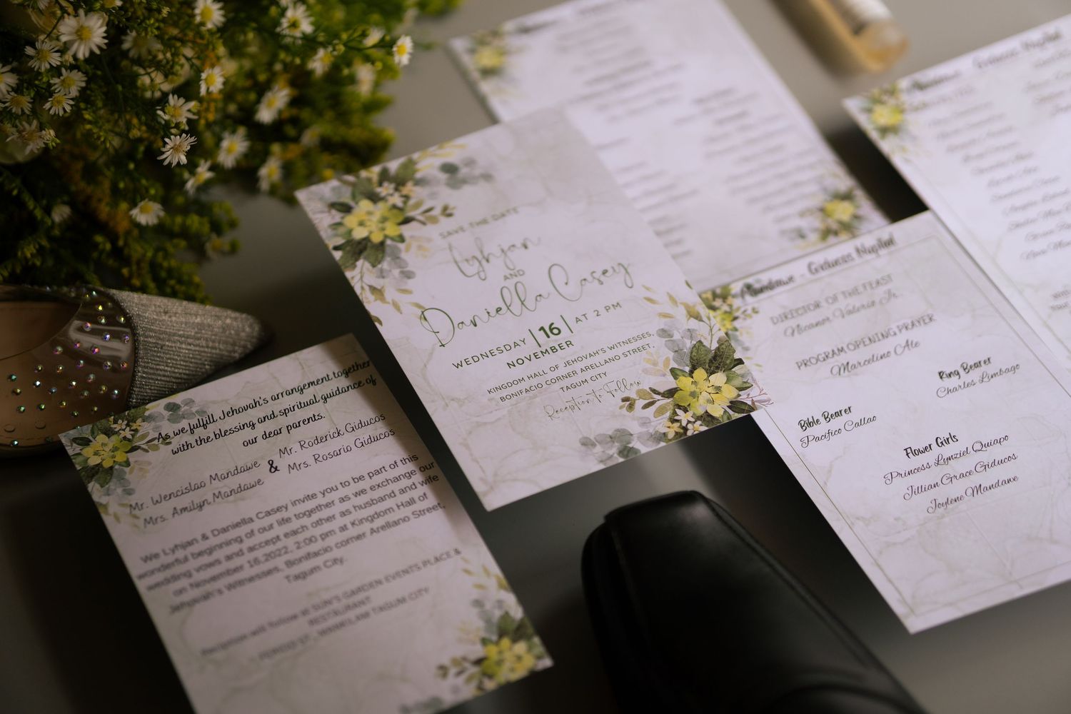 Elegant wedding stationery with yellow floral accents displayed on a dark surface beside a potted plant and bridal shoes.