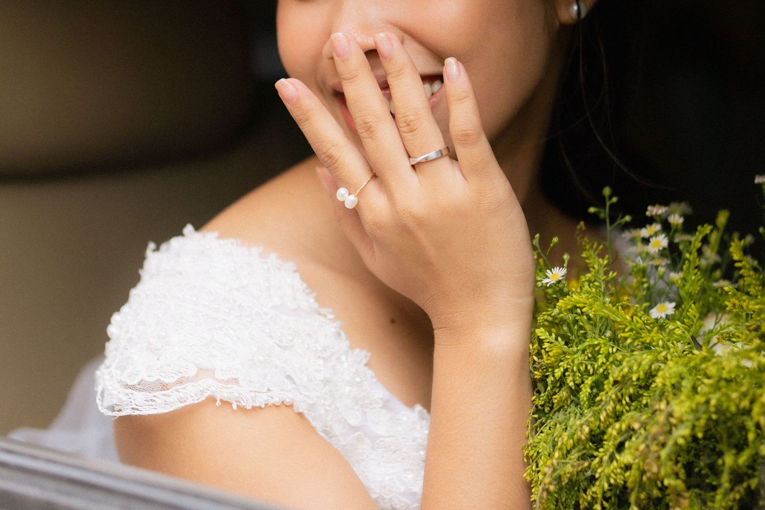 Bride in off-shoulder lace dress covering smile, showing pearl engagement ring, holding green bouquet.