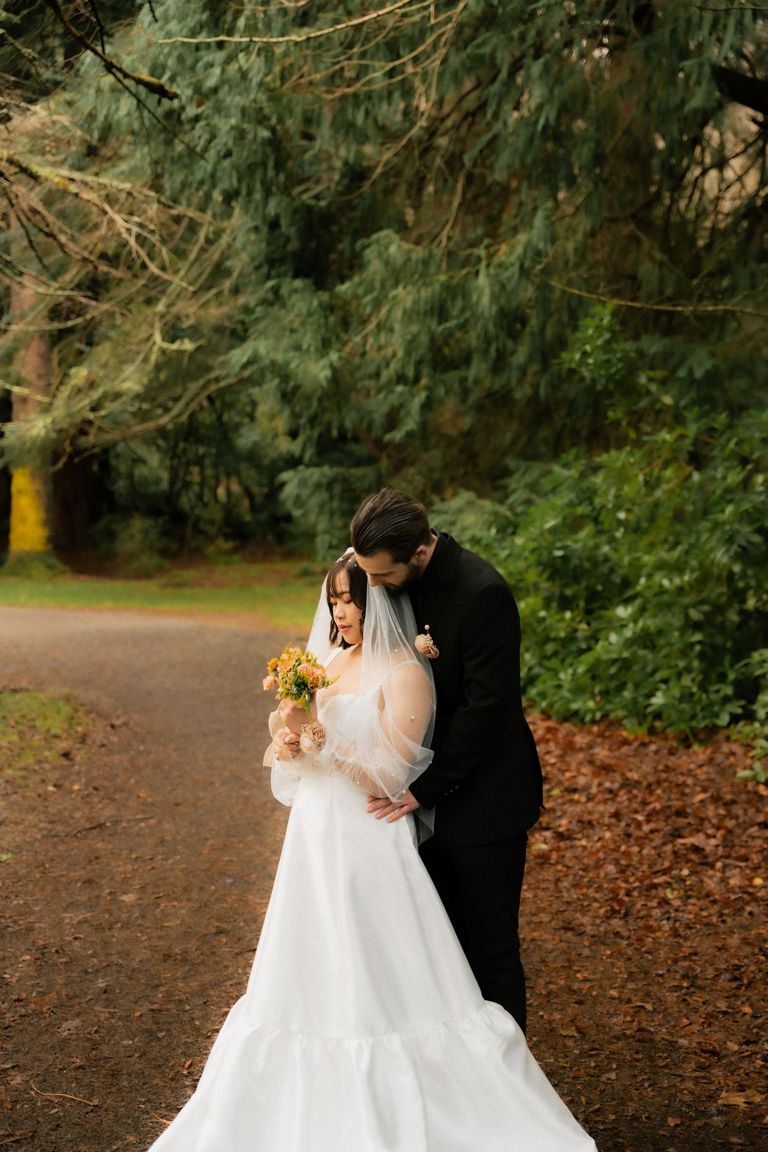 Groom in black suit kissing bride in white gown and veil holding yellow bouquet on a tree-lined path.
