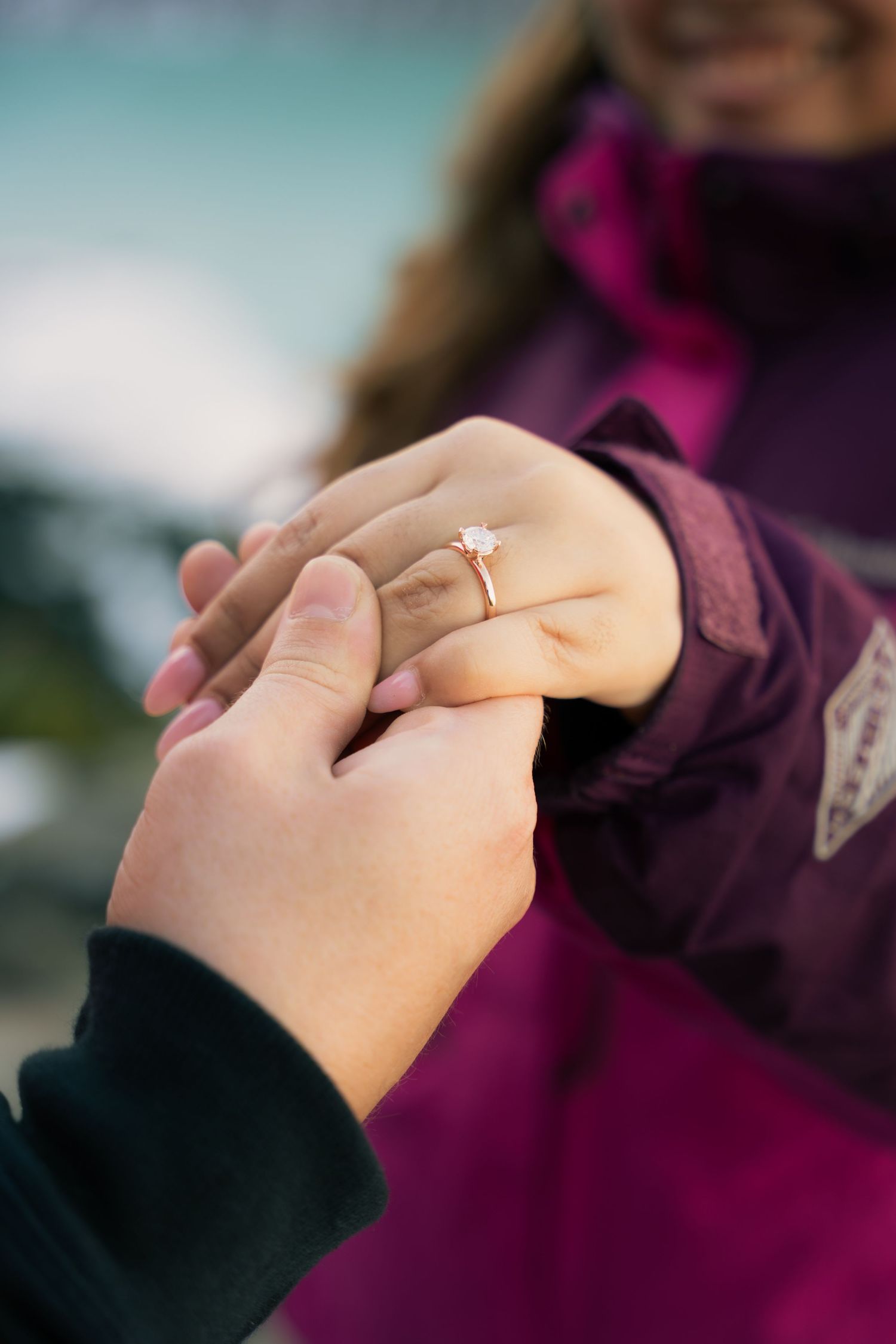 Close-up of a proposal moment with a gold diamond engagement ring being placed on a woman's finger outdoors.