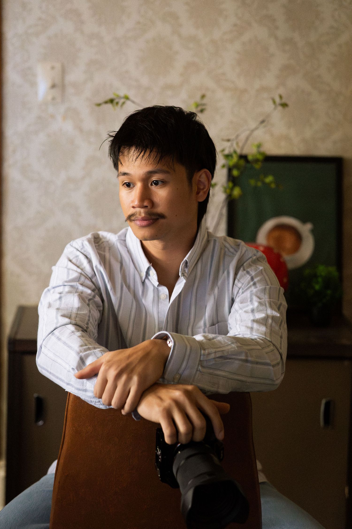 Young Asian man with mustache sits relaxed holding a camera, wearing a striped shirt in a warmly lit vintage room.