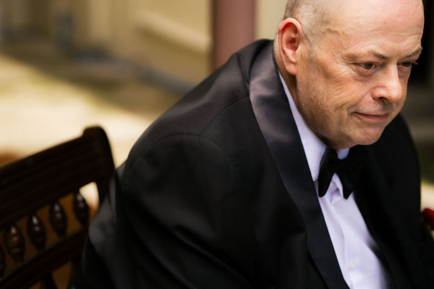 Elderly man in black tuxedo and bow tie leaning forward thoughtfully at a formal event.
