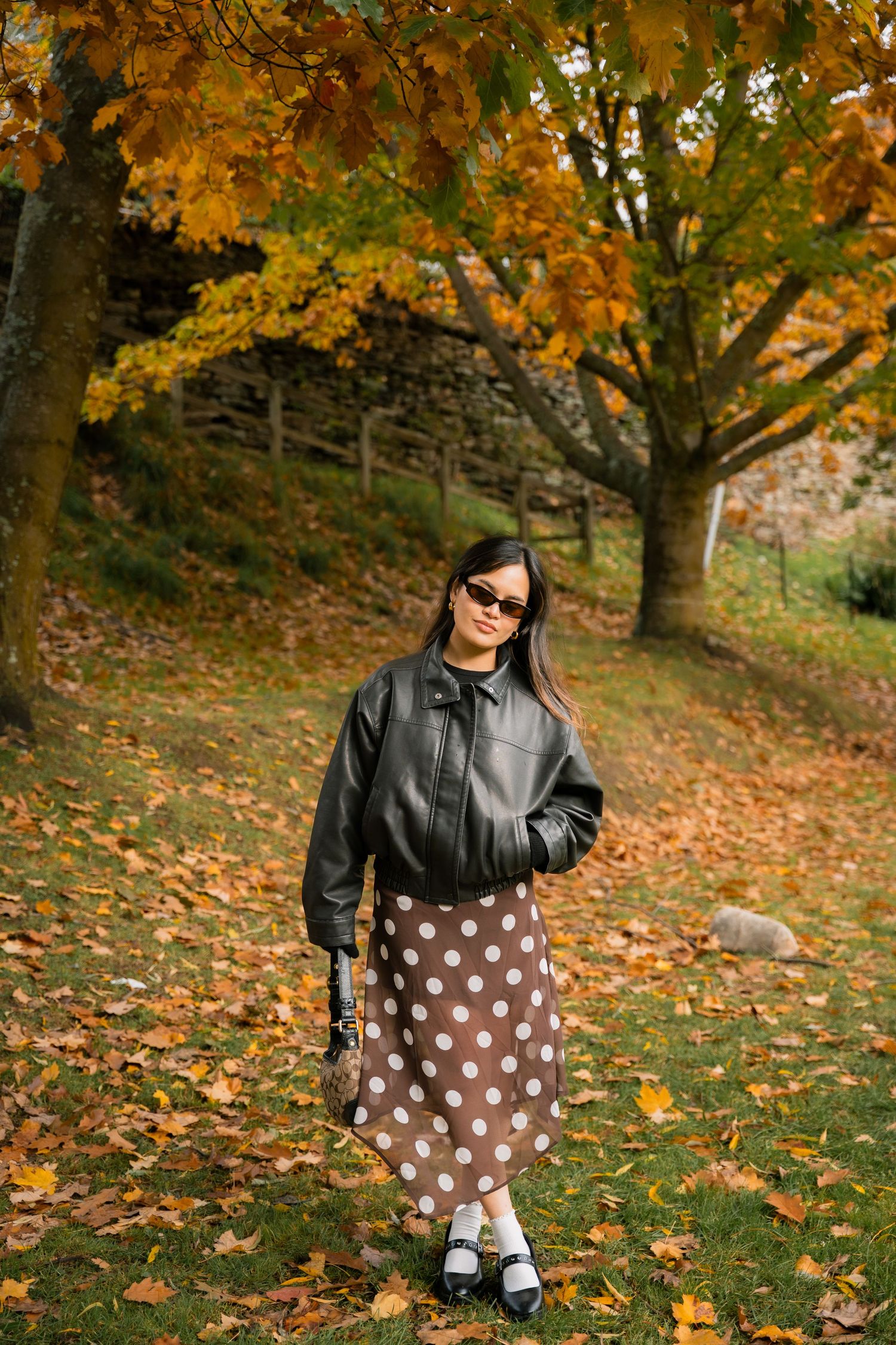 Woman in black leather jacket and polka dot skirt standing among autumn leaves in a park.