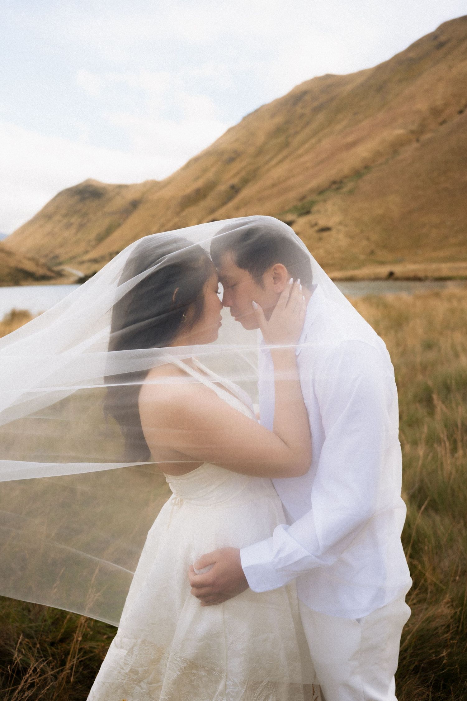 Bride and groom share romantic moment under wedding veil with scenic mountain lake backdrop.