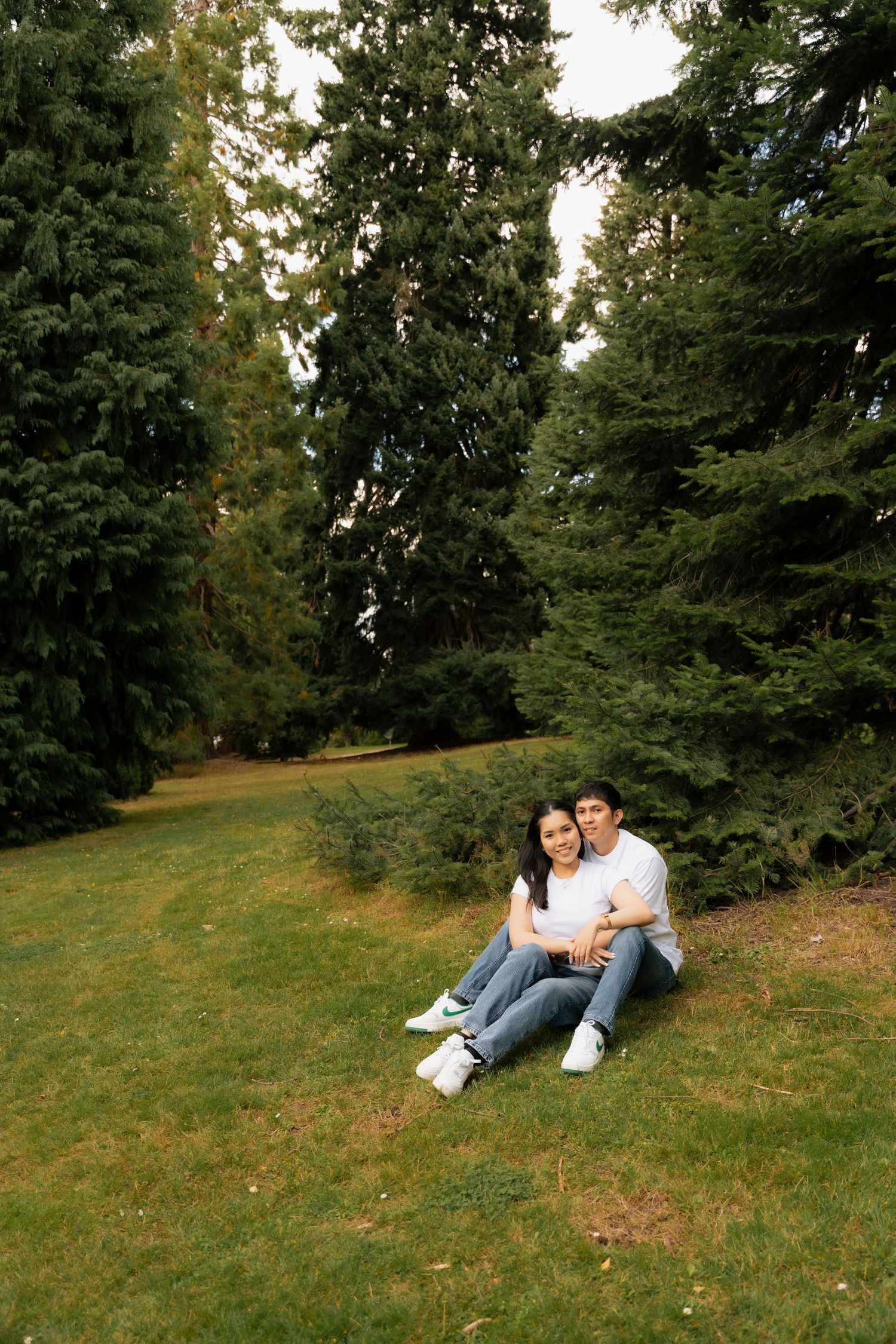 Happy couple sitting together on green grass in a lush park surrounded by tall evergreen trees.