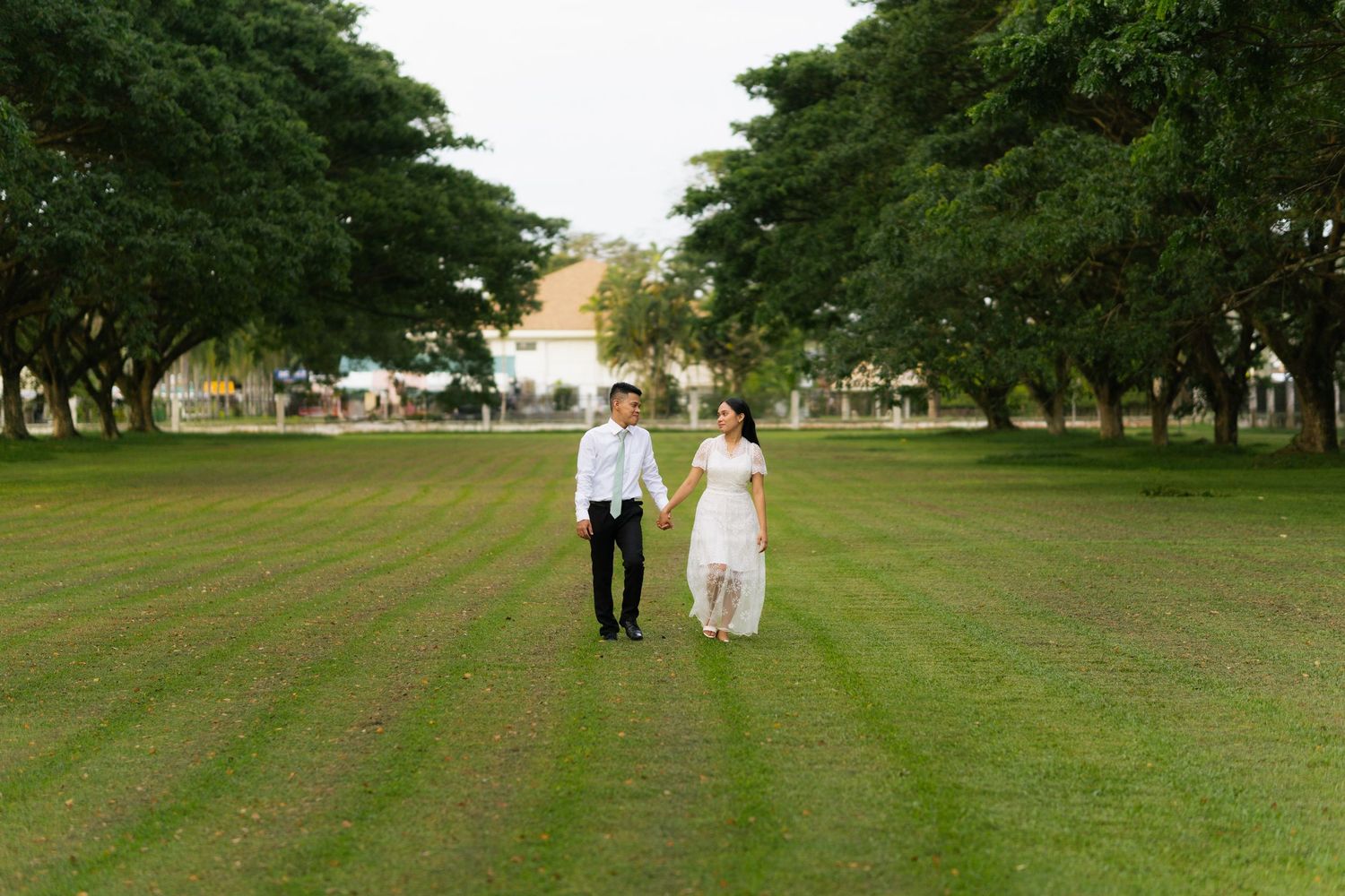 A couple in formal attire walks hand in hand across a wide green lawn lined with tall trees.