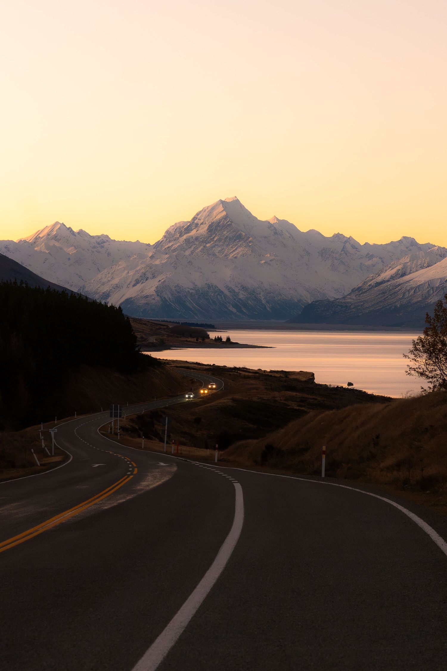 Winding road leading to snow-capped Mount Cook at sunset, with Lake Pukaki reflecting golden hues in New Zealand.