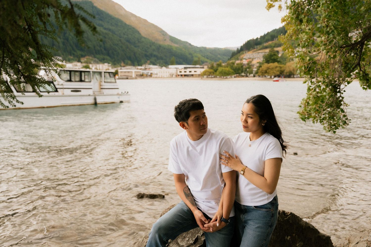 Couple in matching white t-shirts and jeans sitting on rocks by a lake with mountains and boats in the background.