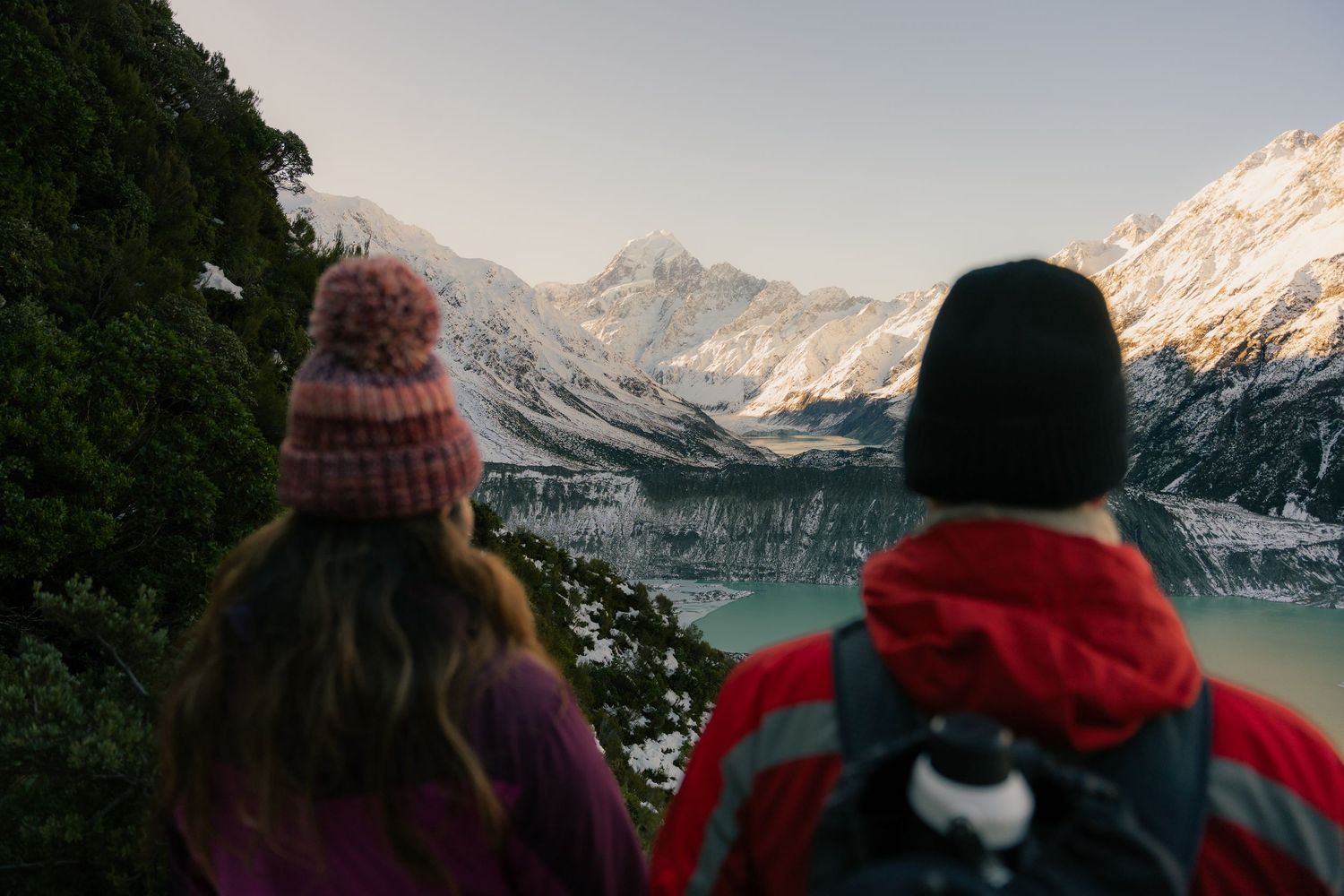 Two people viewing snow-covered mountains and turquoise glacial lake from a forested hiking trail.