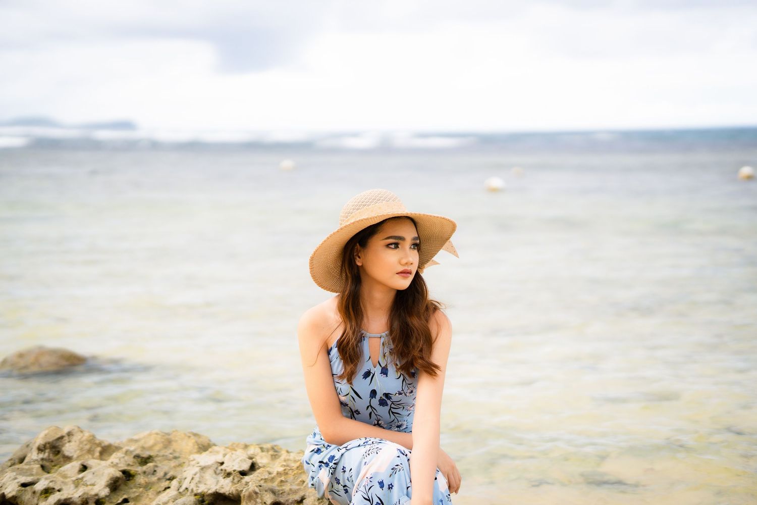 Young woman in floral dress and sun hat sitting on rocks by the ocean on a cloudy day.