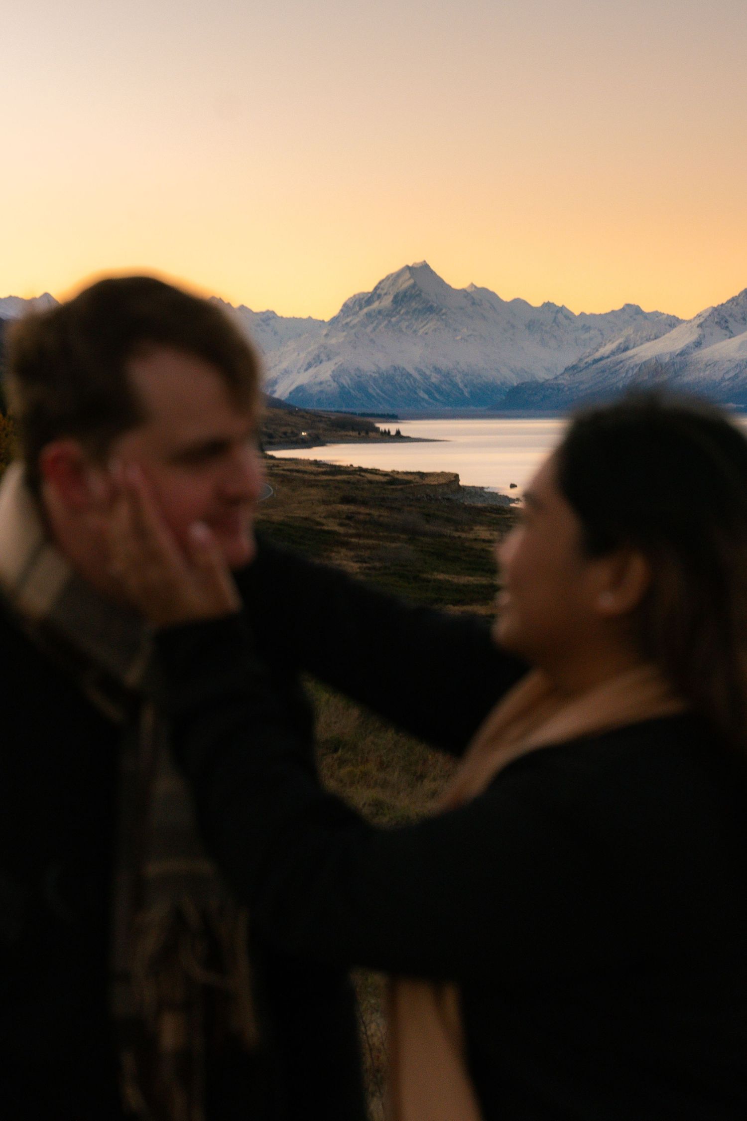 Couple sharing tender moment at dusk with iconic mountain peak and lake visible in the background.