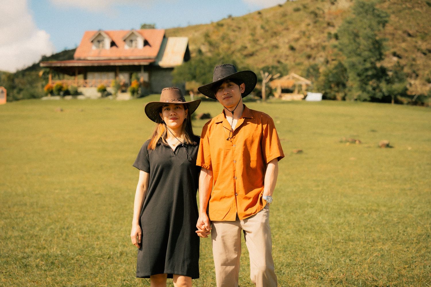 Couple in wide-brim hats holding hands on green lawn with rustic house and hills in background.