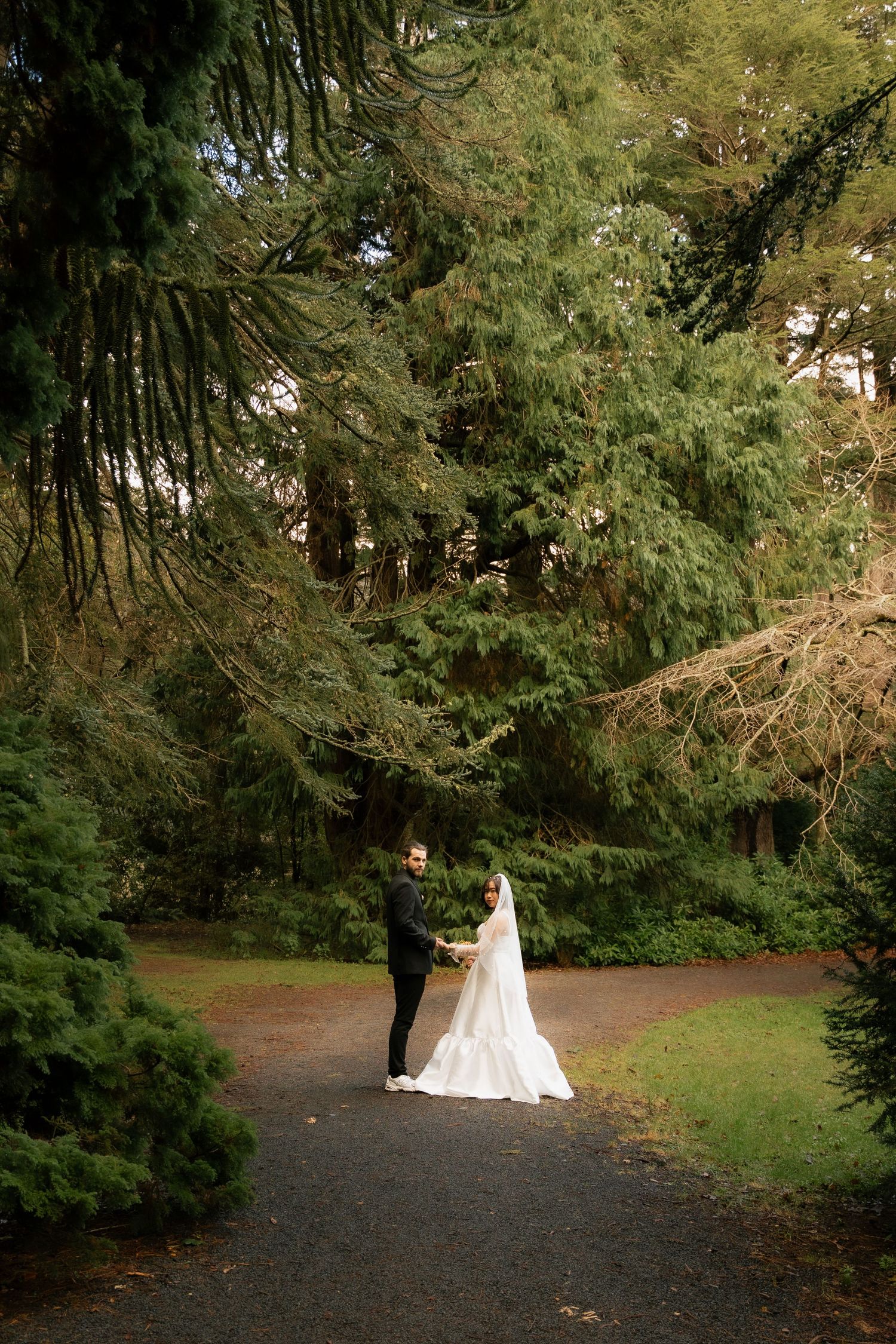 Bride and groom holding hands on a garden path surrounded by lush green trees in an outdoor wedding portrait.