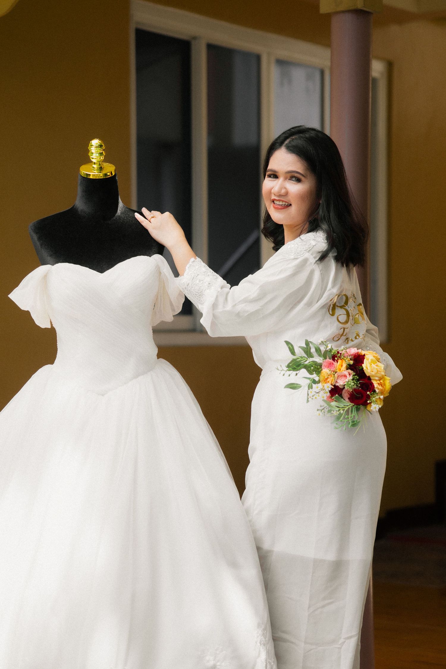Woman in white dress holding colorful bouquet stands beside a wedding gown on a mannequin indoors.