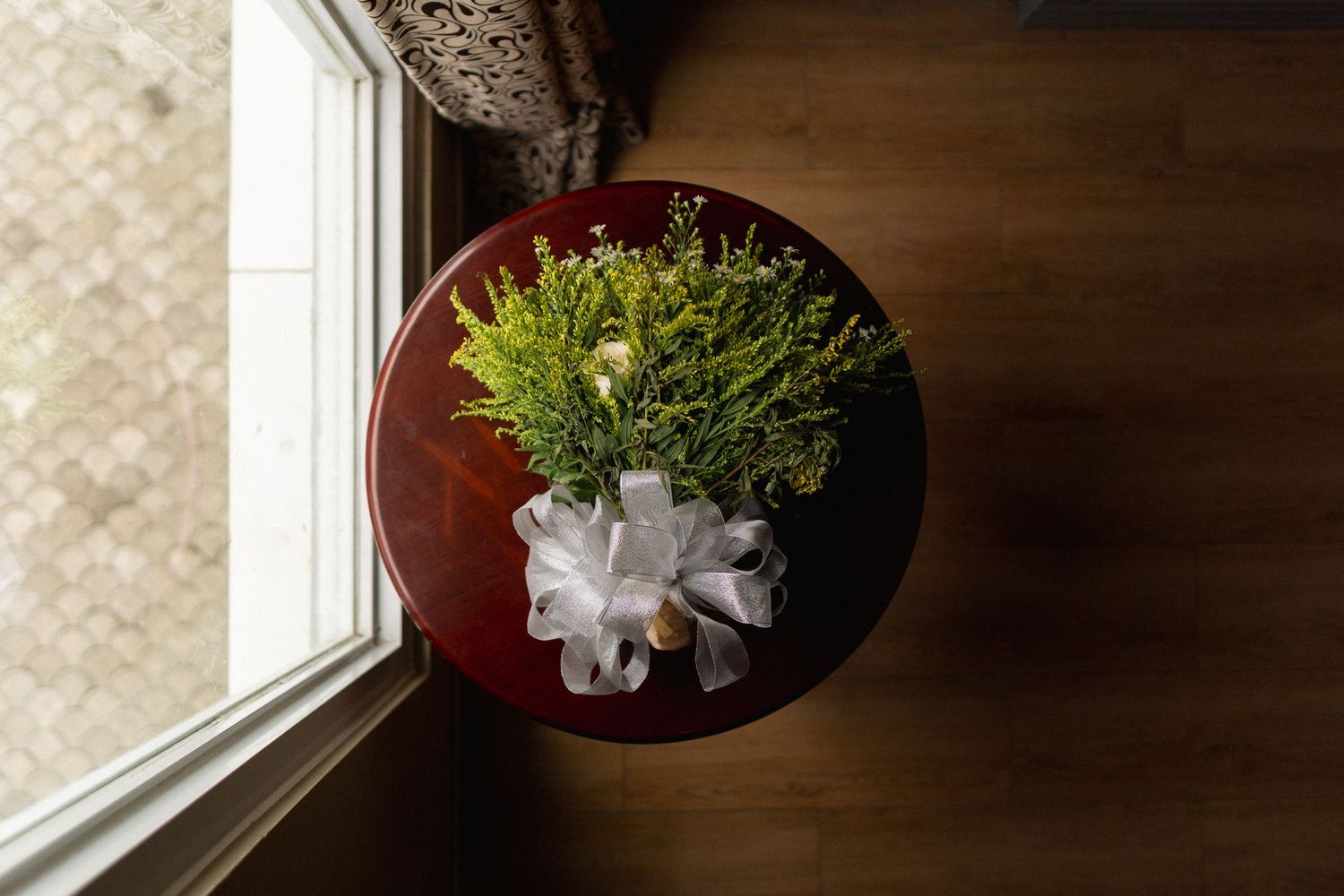 A small round wooden table by a window holds a bouquet of yellow flowers wrapped in white ribbon.