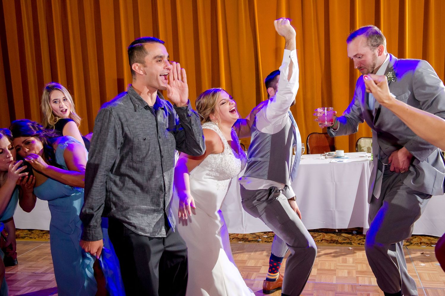 Wedding party dances energetically under orange and purple lighting at reception.