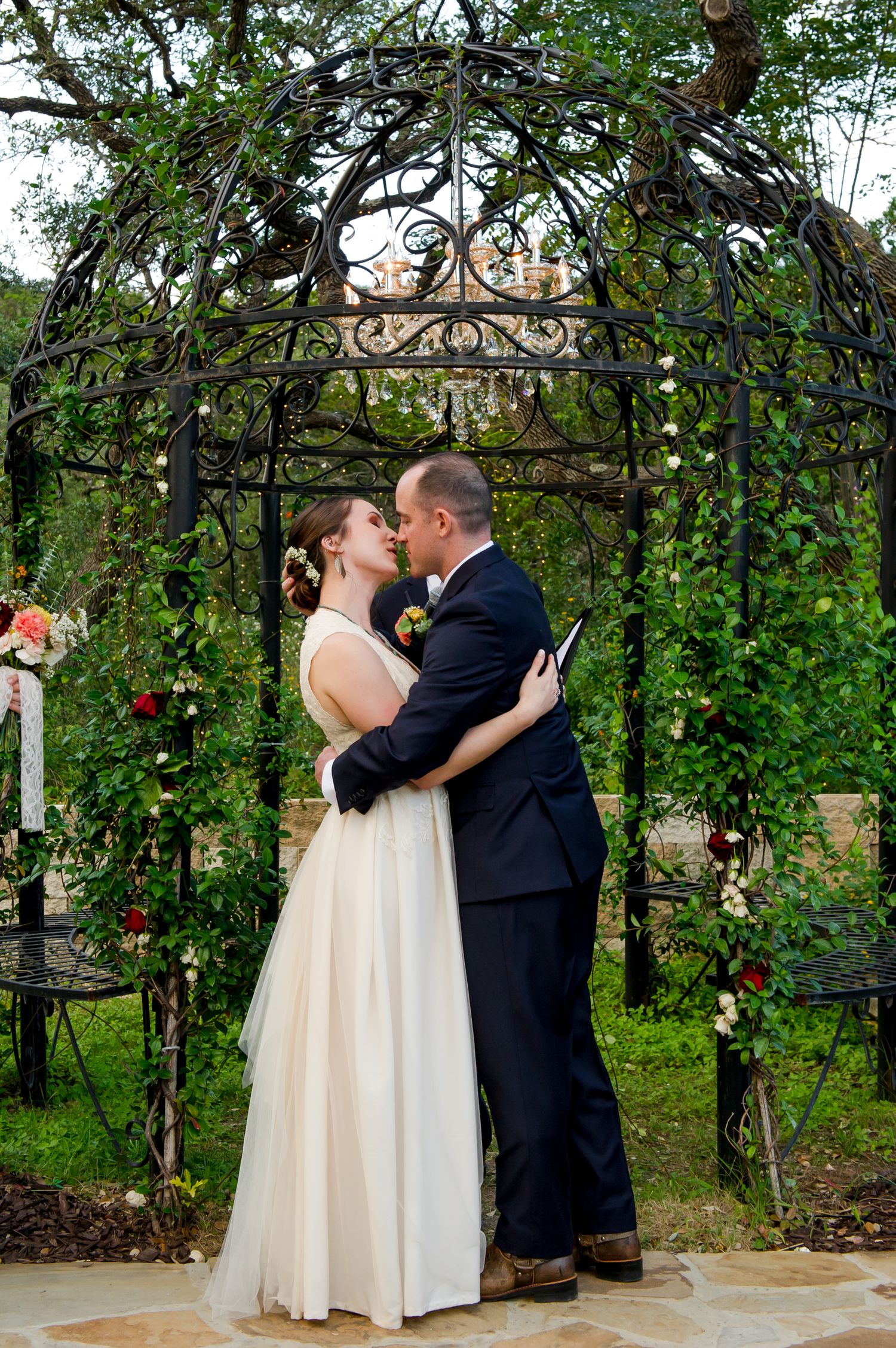 Romantic outdoor wedding ceremony under a floral arch with trees and greenery in the background.