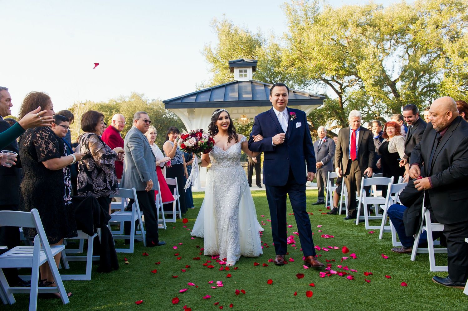 Outdoor wedding ceremony setup with white chairs and rose petals on green lawn.