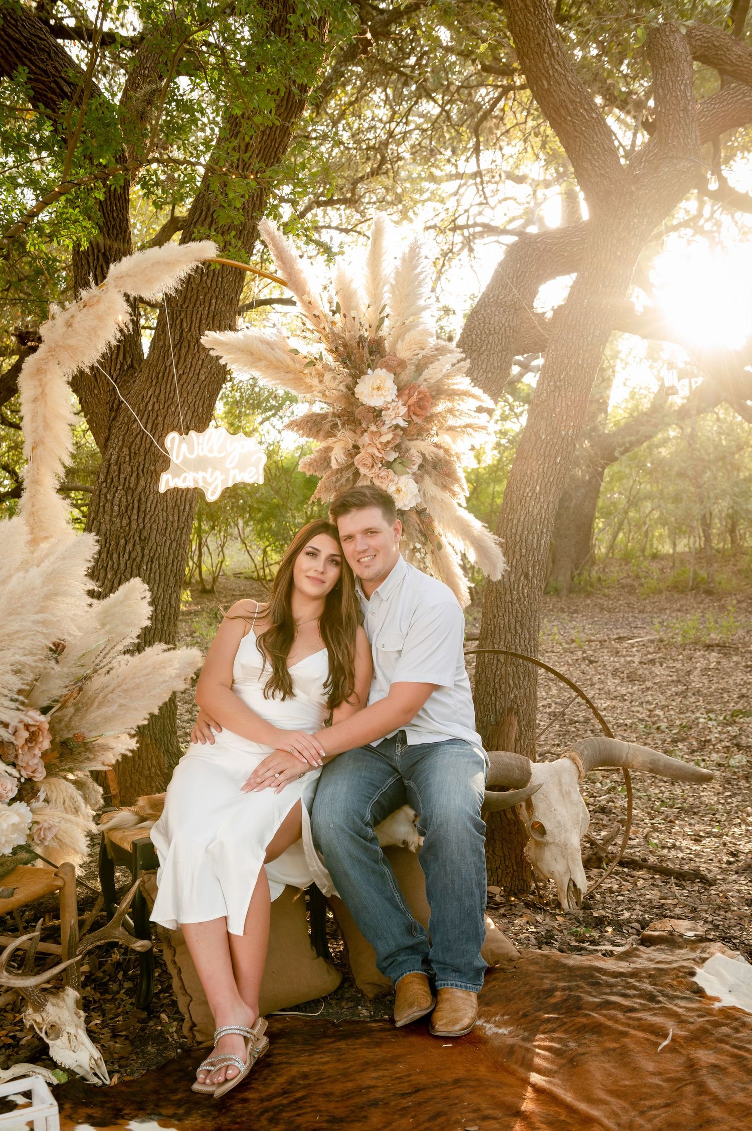 Couple sitting on vintage furniture with boho dried flower arch in a rustic outdoor setting at golden hour.